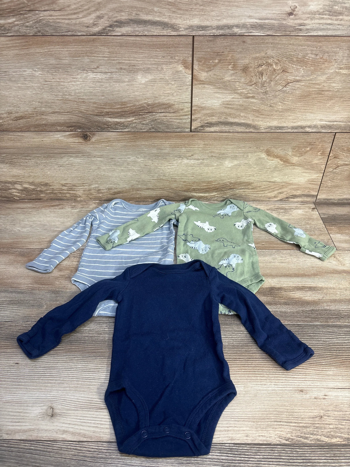 Front view of three Carter's long-sleeve baby bodysuits (size 3M) showing navy solid, gray stripe, and sage dinosaur print on wood background