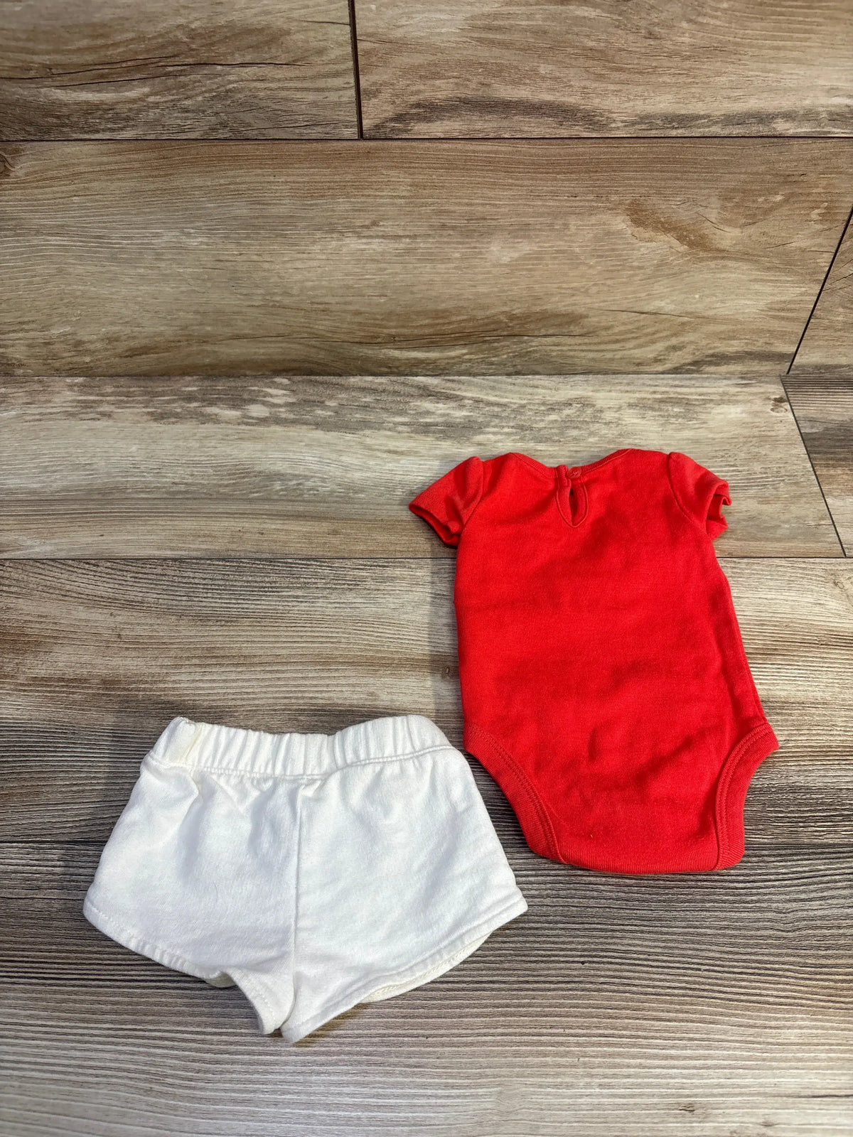 Overhead view of Carter’s 2-piece shirt and shorts set showing a red bodysuit and white shorts on a wood-look floor