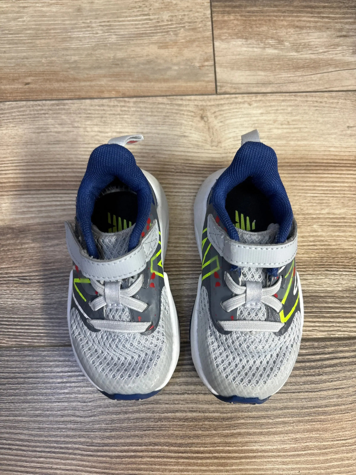 Top-down view of New Balance Rave Run v2 toddler sneakers, white/blue, showing mesh upper, neon accents and velcro strap on wood floor