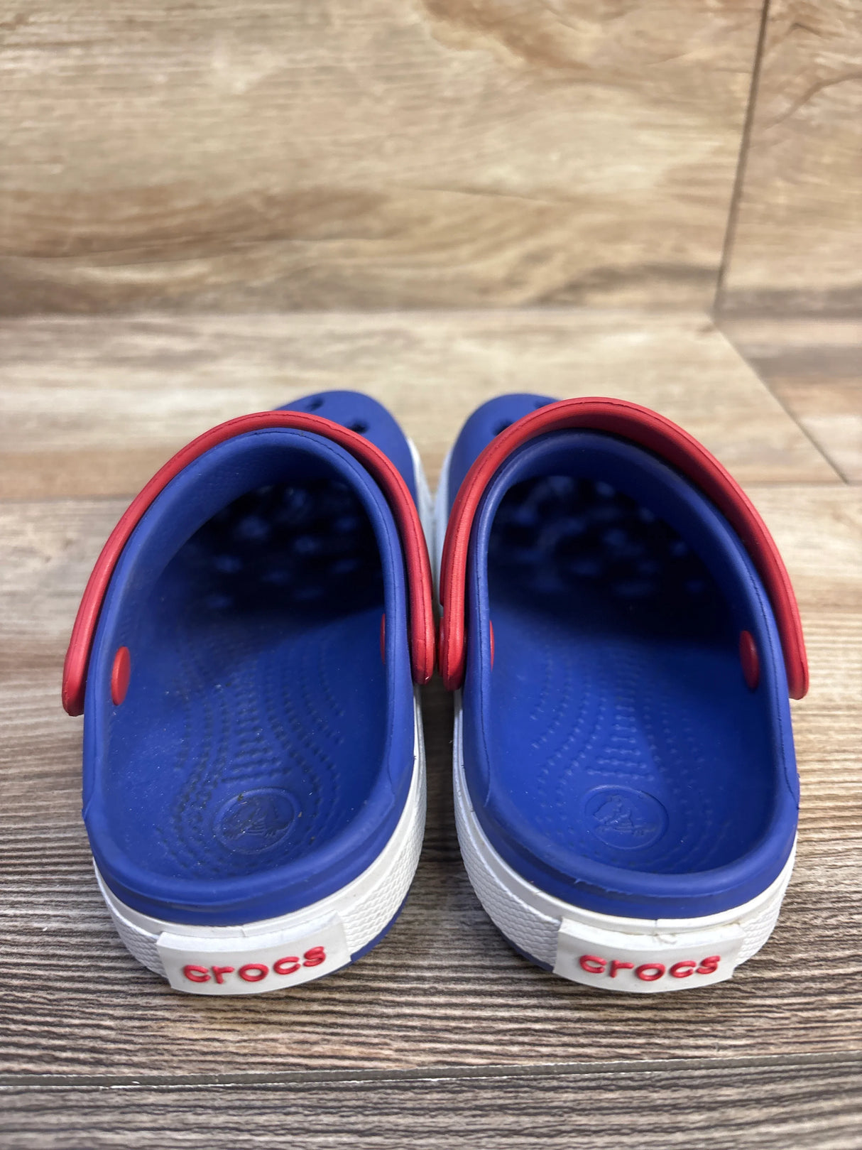 Rear view of Crocs Crocband II kids’ blue clogs showcasing red heel straps and white soles on wood floor