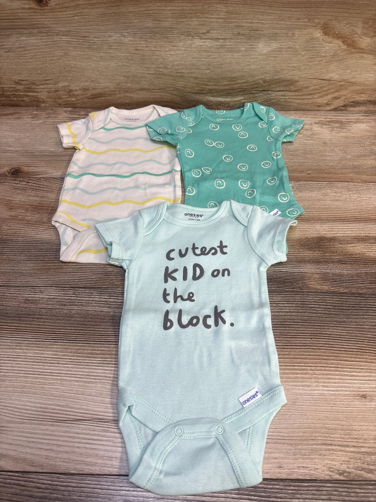 Top-down of three Onesies bodysuits: mint with text cutest KID on the block, teal smiley print, and white striped on wood