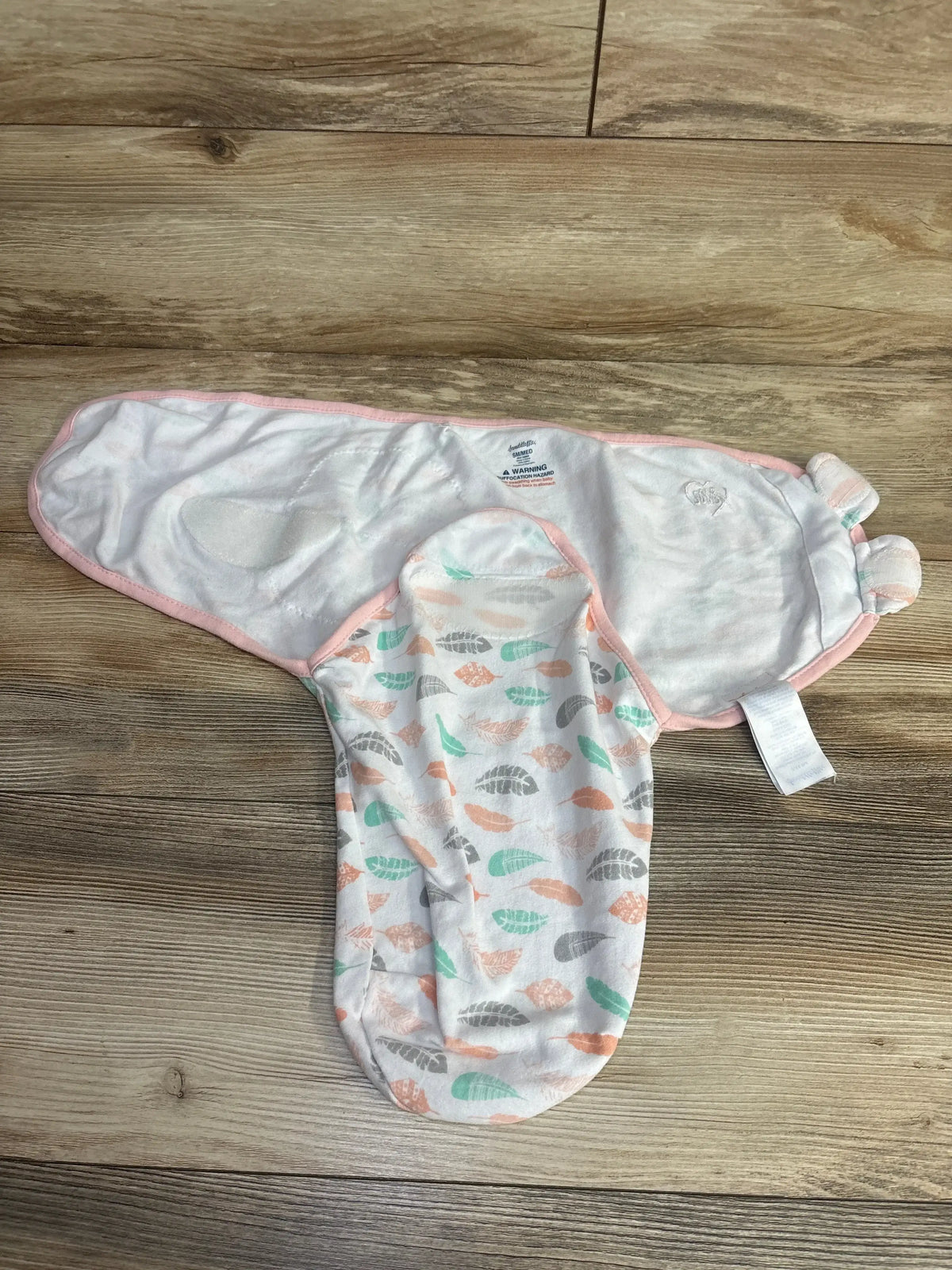 White and pastel feather patterned baby swaddle wrap with pink trim on wooden floor