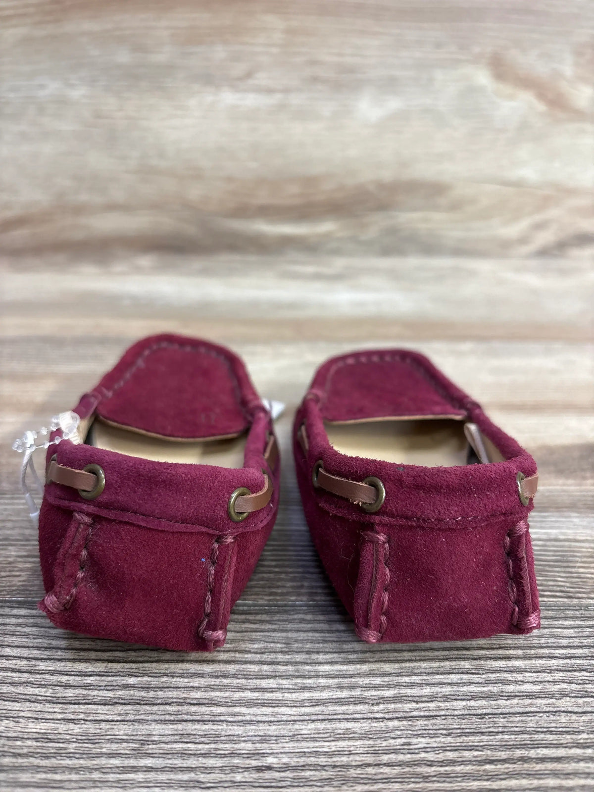 Pair of burgundy suede baby moccasin shoes with brown leather lace detail on wooden surface