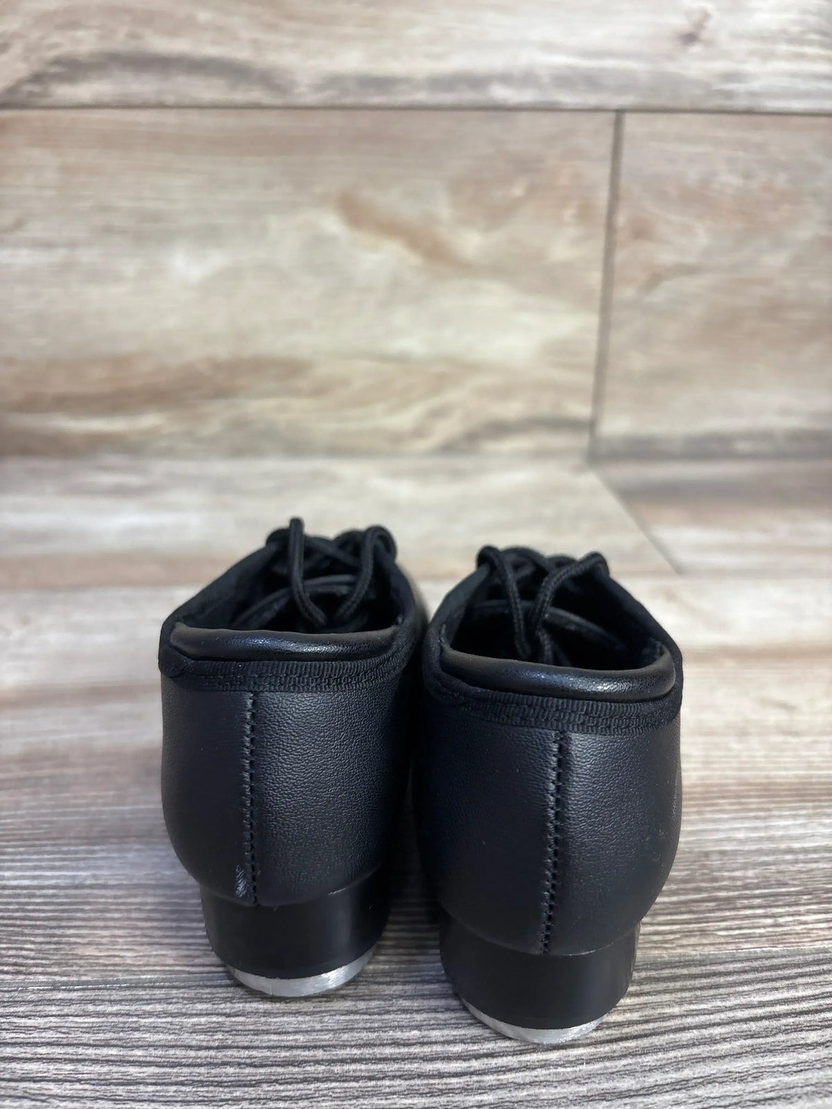 Rear view of Theatricals black tap shoes showing stitched heels, laces, and metal heel taps on wood floor