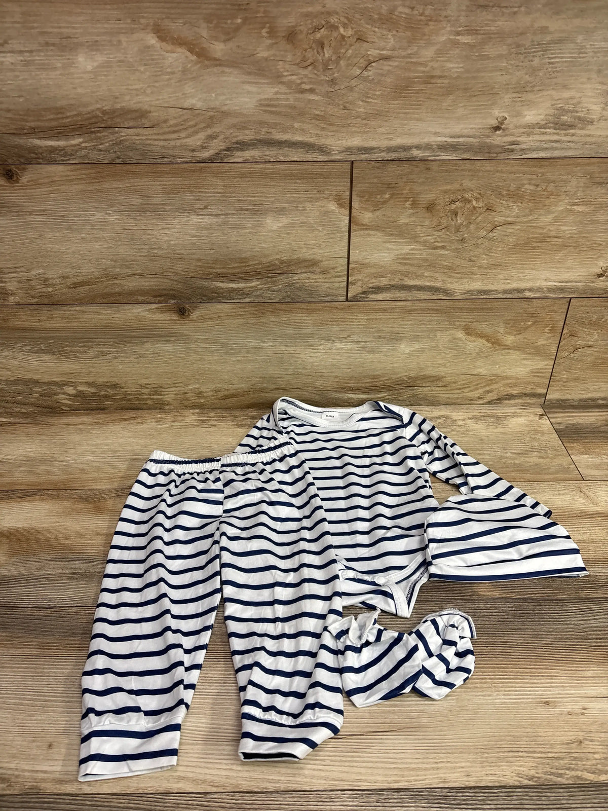 Top-down view of a four-piece baby bodysuit, pants, mittens, and hat set showcasing navy stripes on white fabric with wood background
