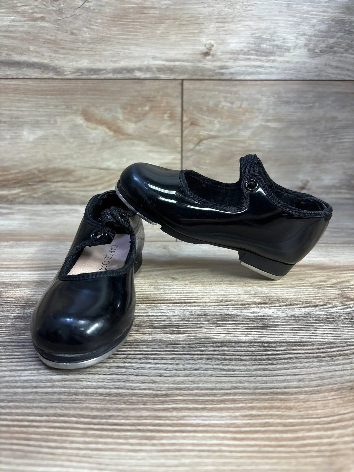 Black patent leather tap shoes with elastic straps on wood floor, Capezio brand