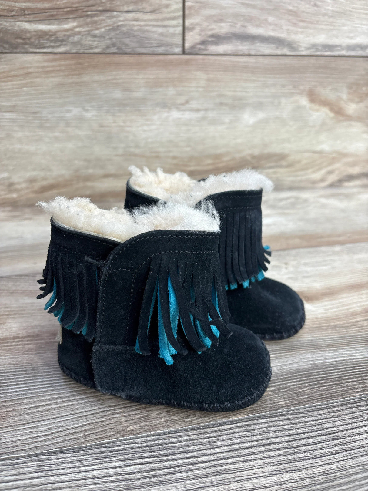 Black suede baby moccasin boots with blue fringe and white shearling lining on wooden surface