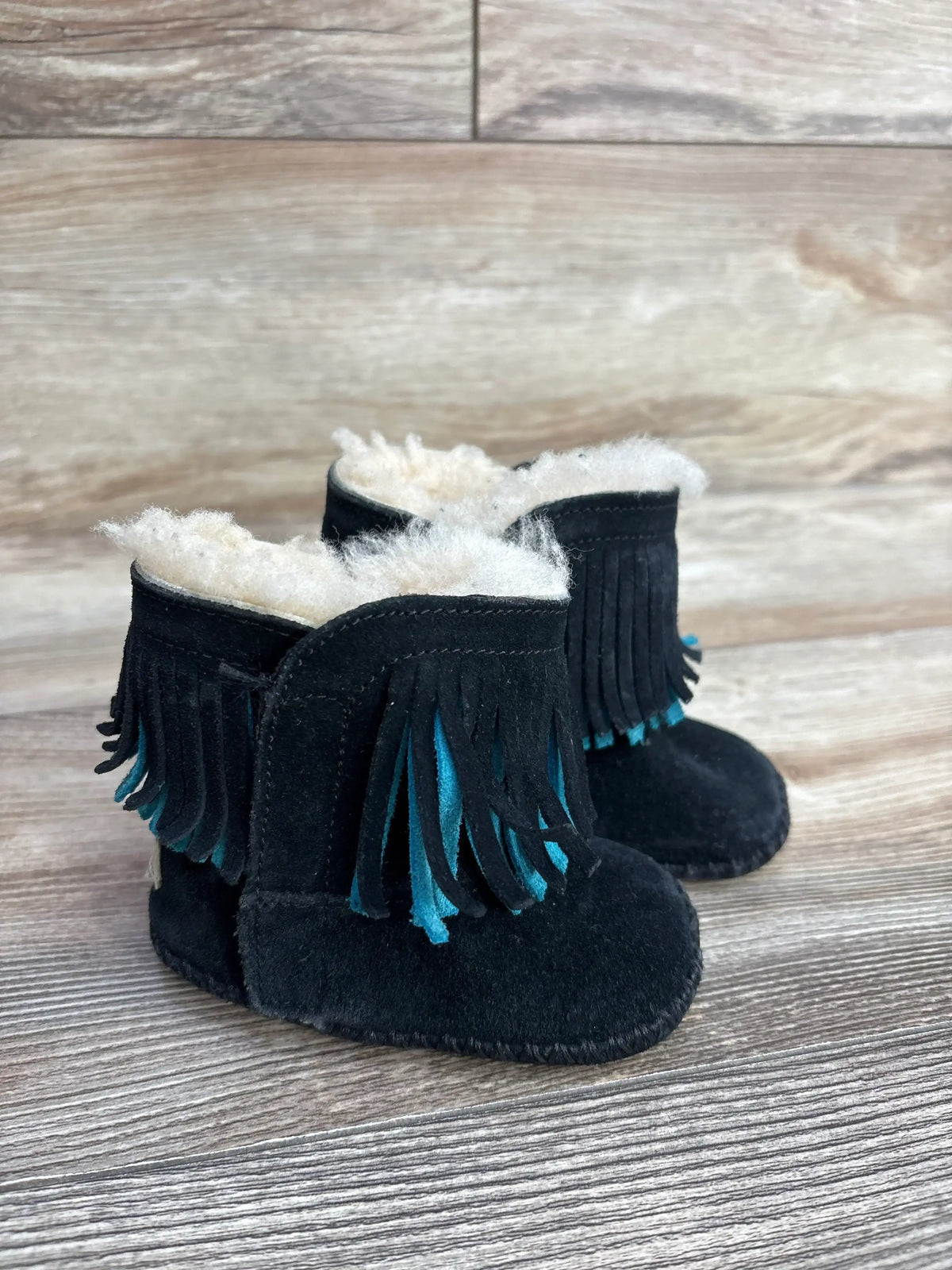 Black suede baby moccasin boots with blue fringe and white shearling lining on wooden surface