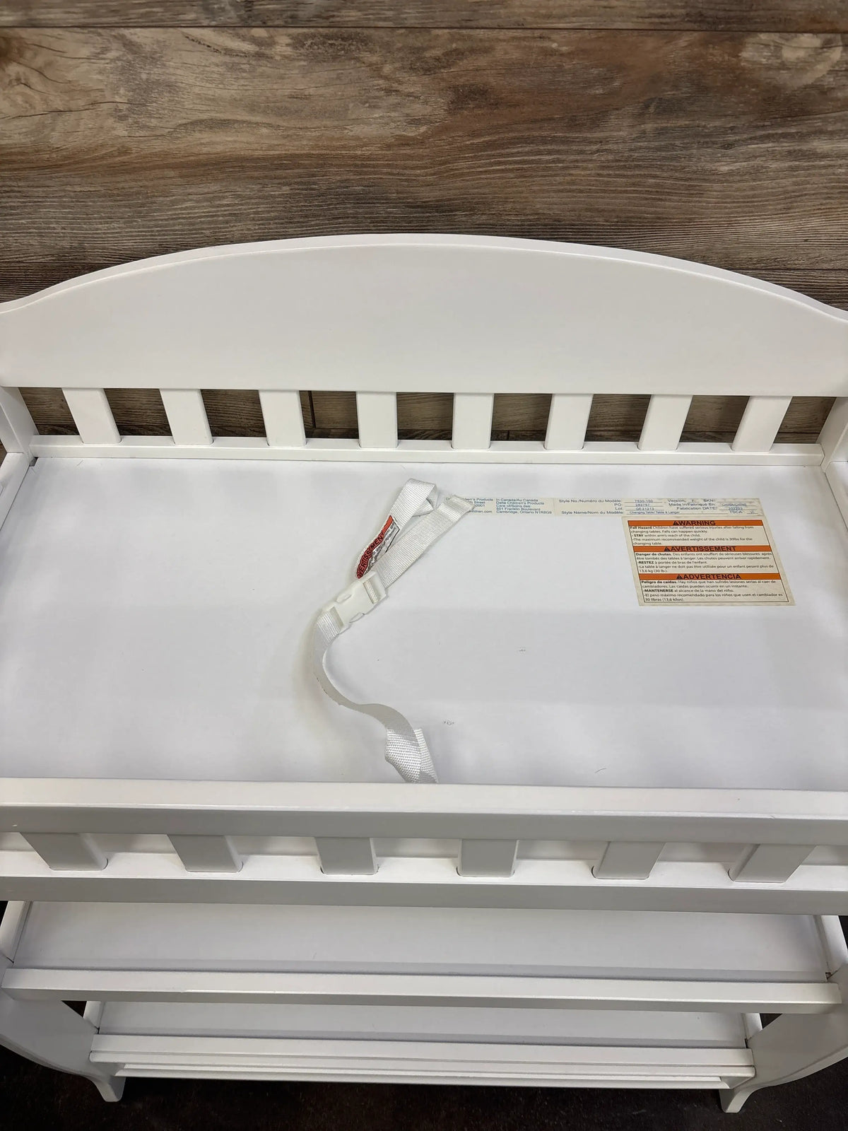 Close-up view of white Delta Children Wilmington changing table showcasing safety strap and open lower shelf with wood backdrop