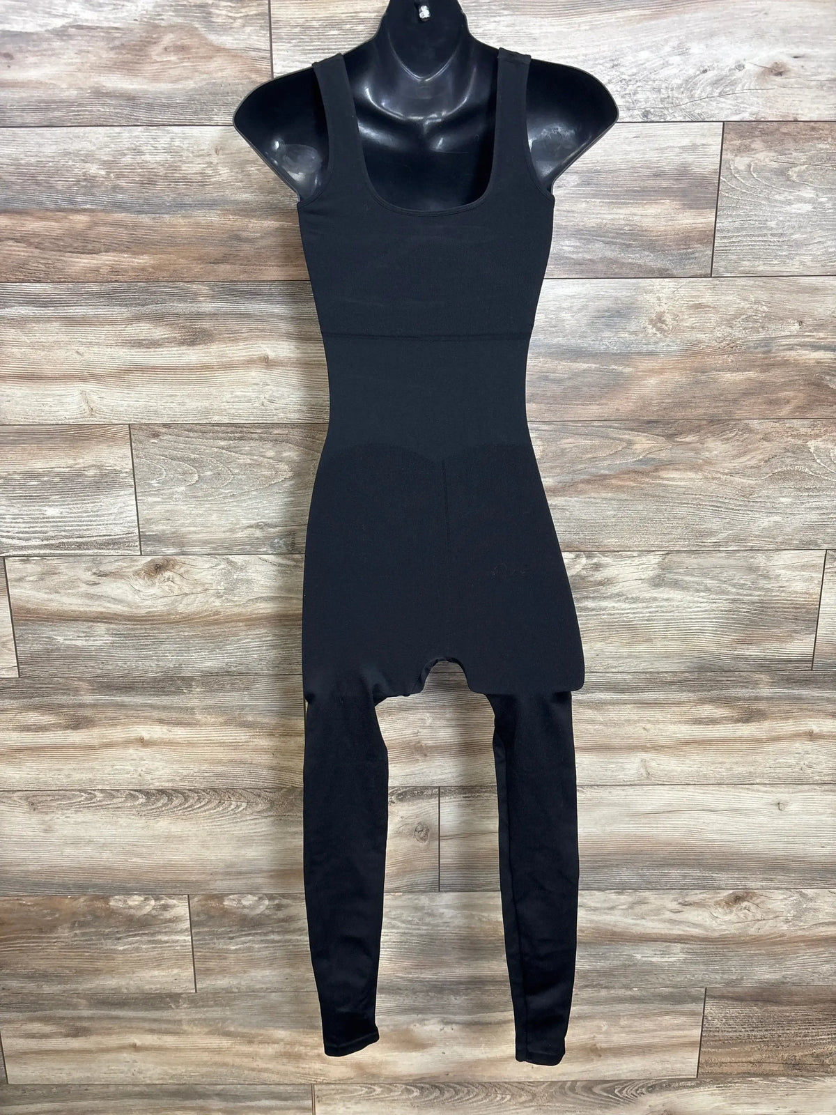 Front view of black maternity compression unitard showcasing wide shoulder straps, high-waist shaping panels, and full-length legs with wood-paneled background