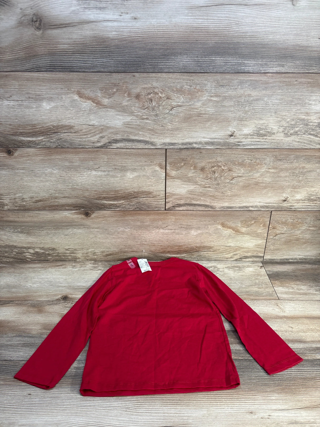 Flat lay of red long sleeve kids shirt showcasing crew neckline and solid color with brand tag on wood plank floor