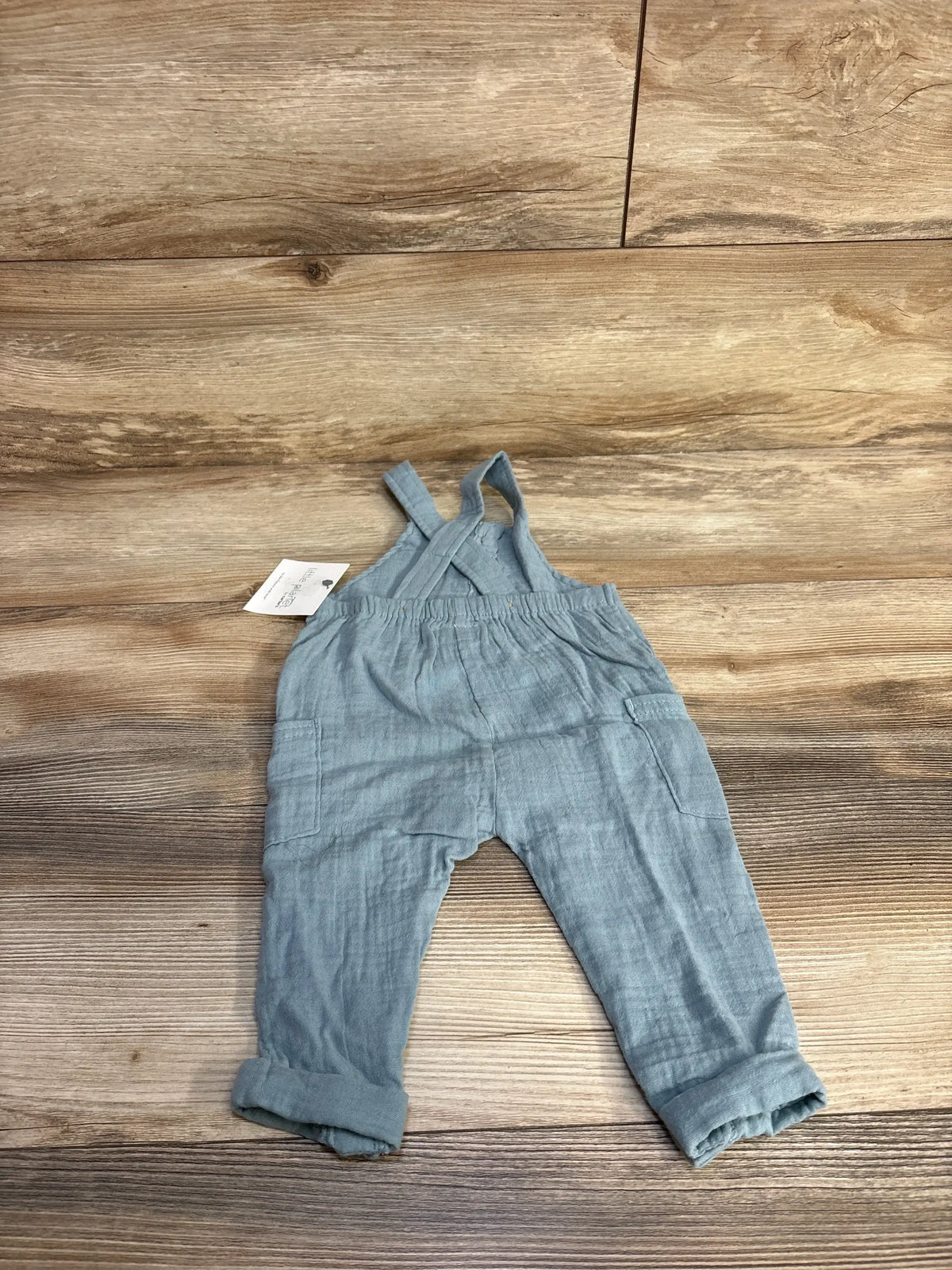 Flat lay Little Planet gauze henley overalls, blue 6M, crossed straps, elastic waist and side patch pockets on wood background