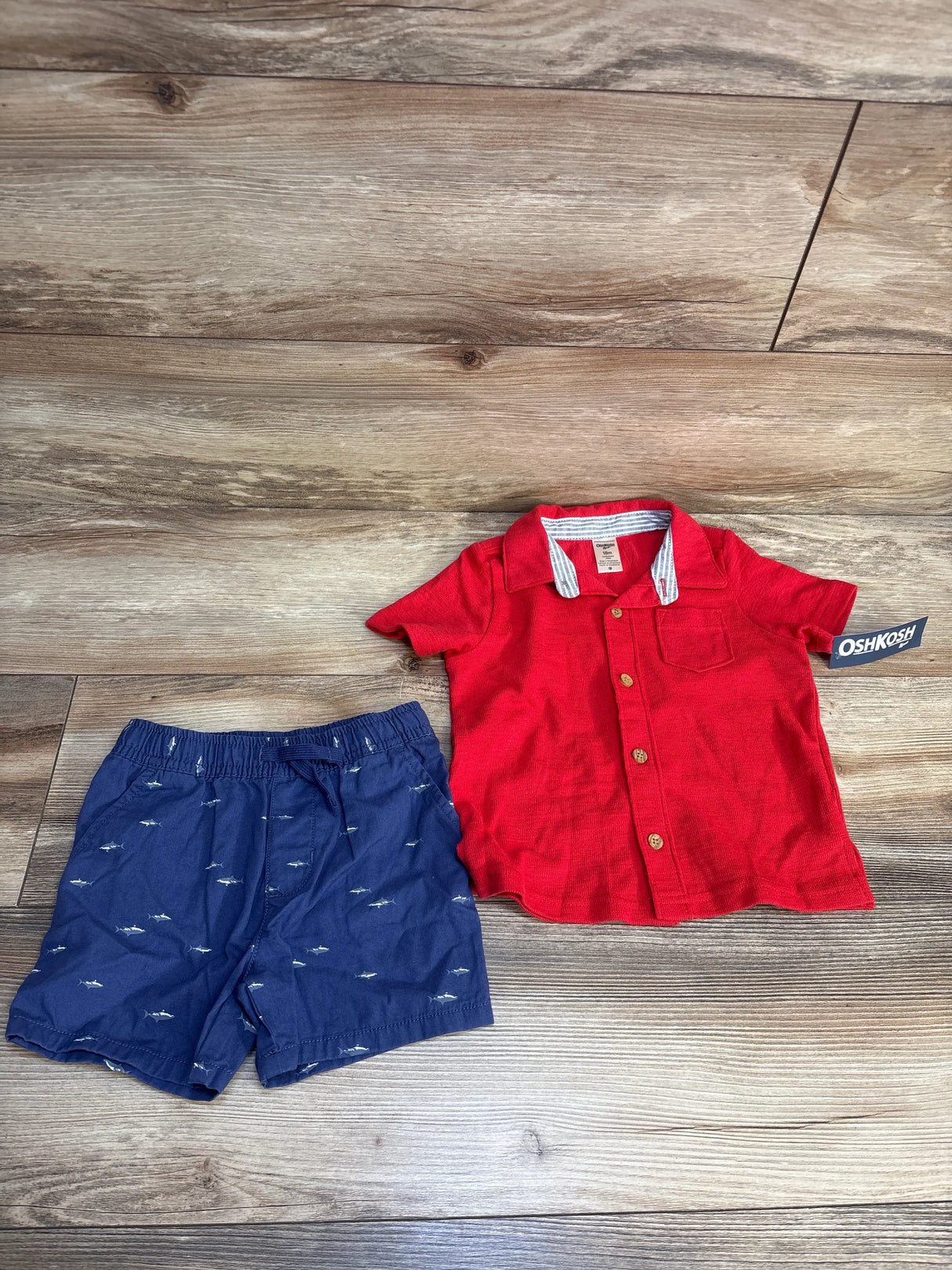 Top view of OshKosh button-down top and woven shorts (18M) with red shirt and navy shark-print shorts on wood floor