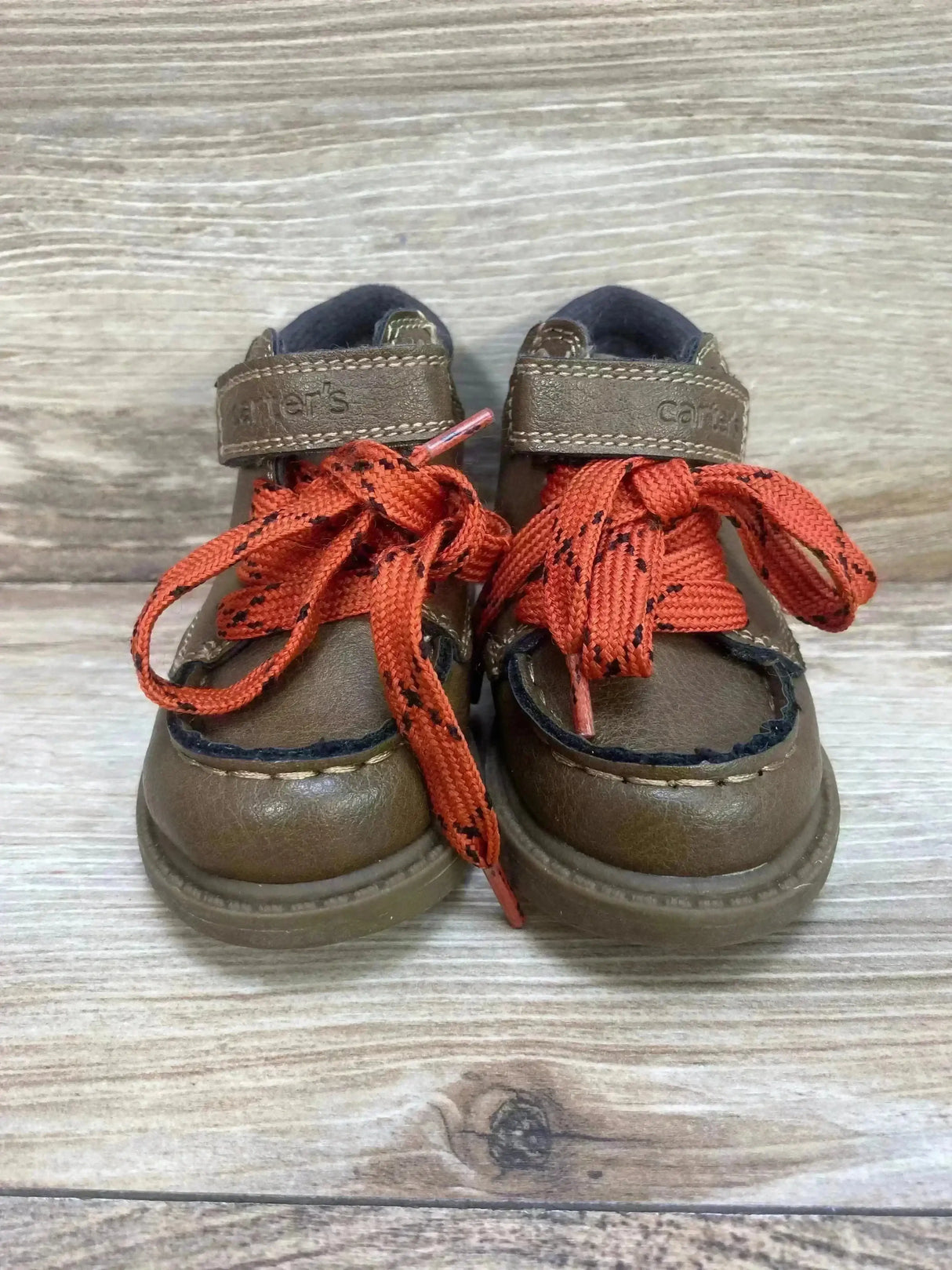 Front view of Carter’s toddler brown Norman ankle boots with orange patterned laces and Velcro straps on light wood background