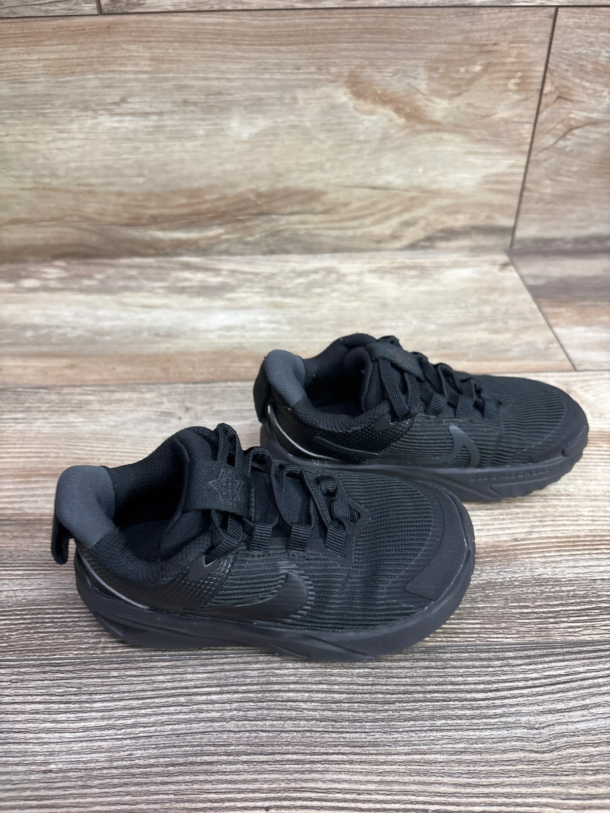 Angled view of Nike Star Runner 4 TD Triple Black sneakers showcasing black mesh uppers and cushioned soles on a wood floor