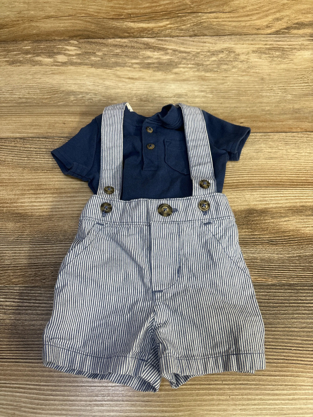 Front view of Just One You newborn two-piece henley shirt and striped suspender shorts on a wood surface