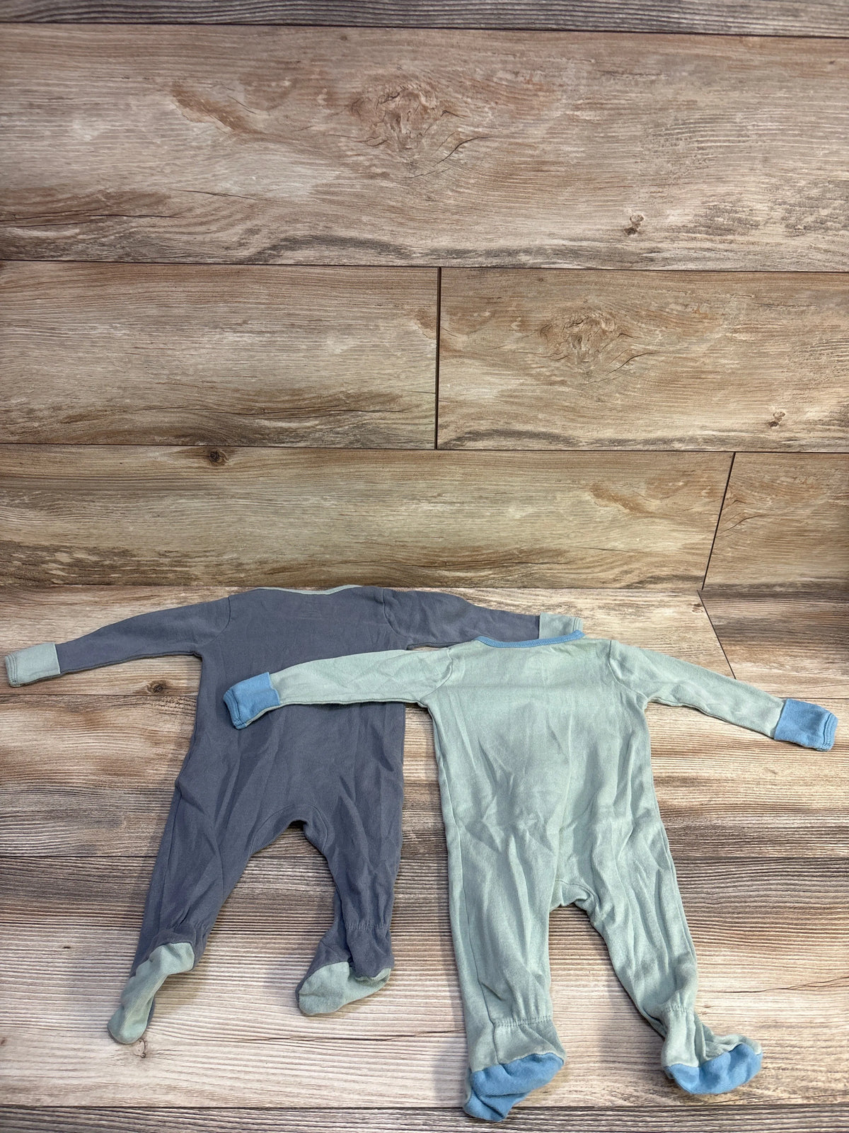 Flat lay of Cloud Island 2-pack footed sleepers (green and grey) showcasing long sleeves and fold-over cuffs on wood background