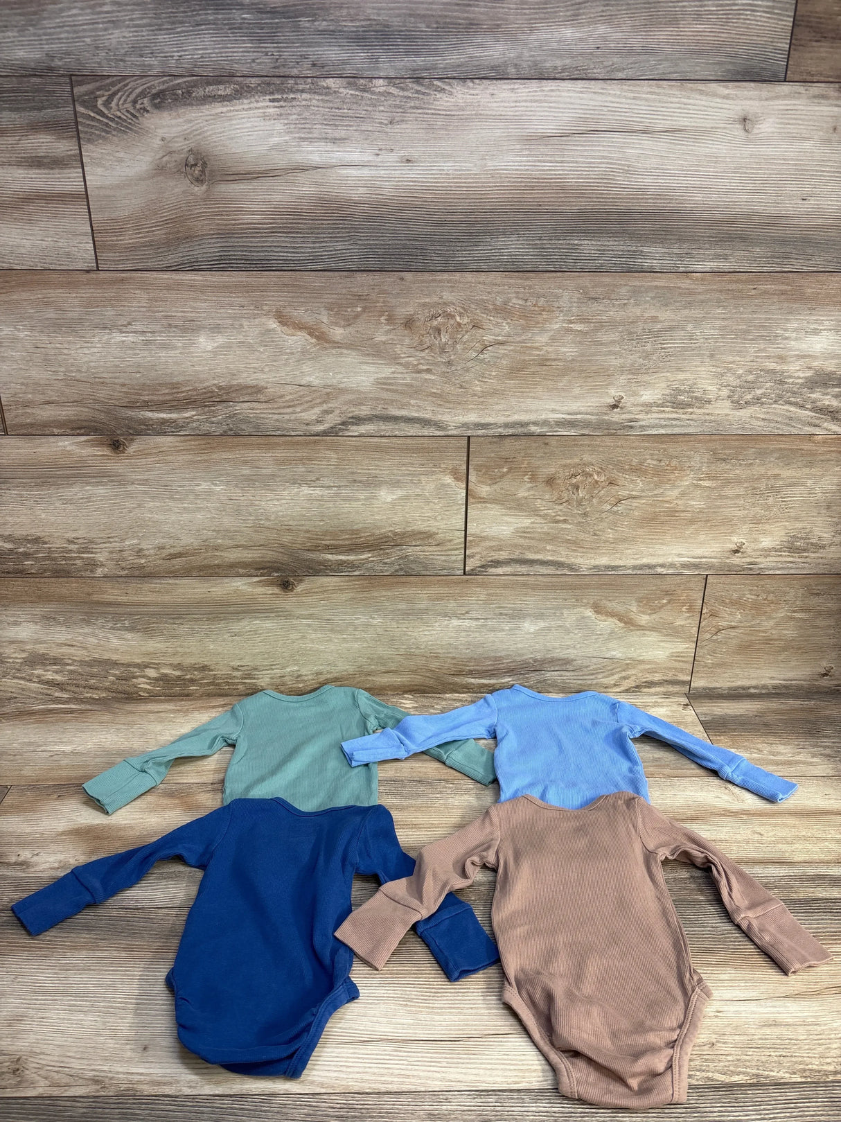 Four long sleeve baby bodysuits in blue, green, brown, and tan on a wood floor