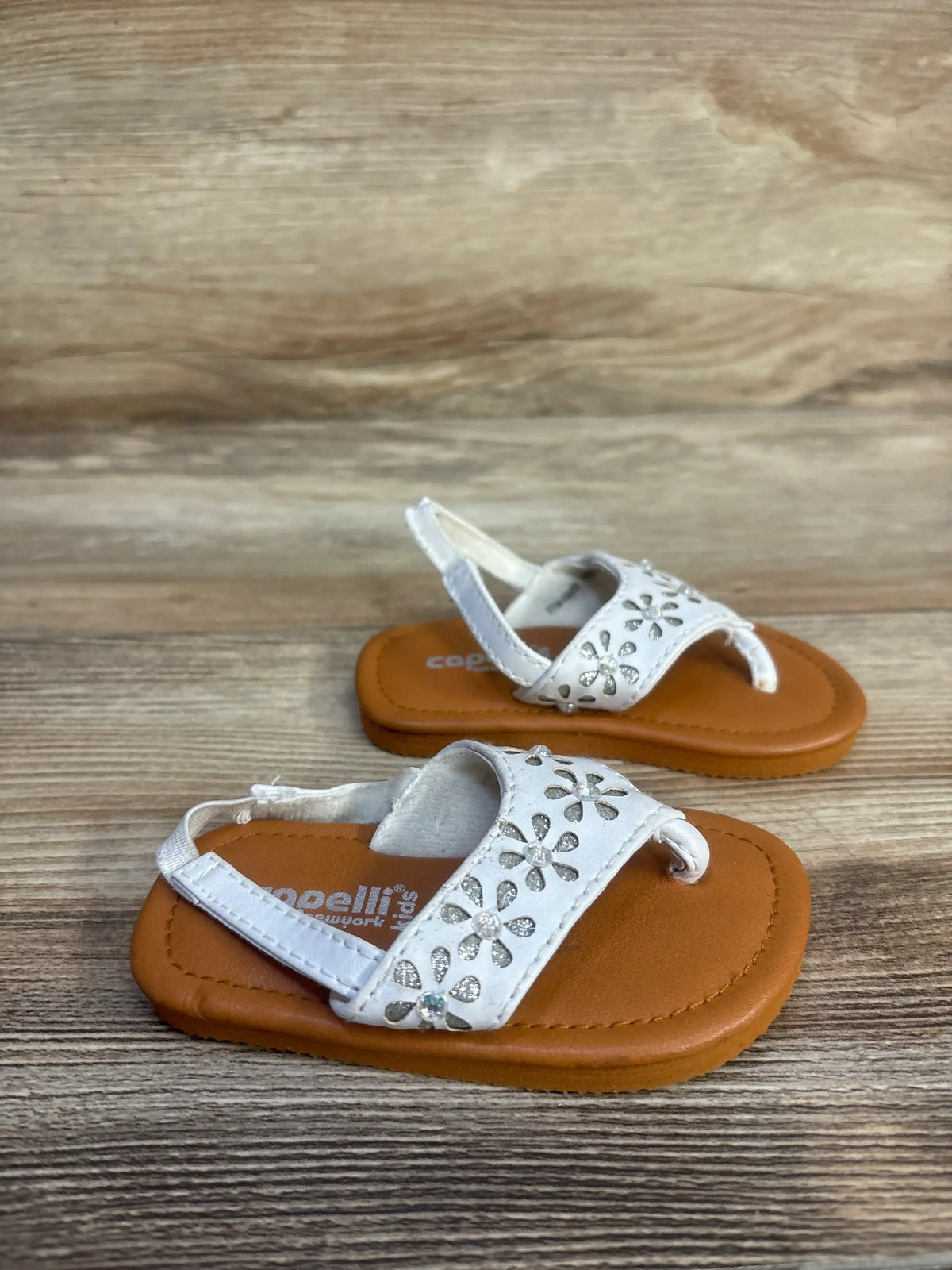 Angled view of Capelli white backstrap sandals, size 4.5C, with floral rhinestone embellishments on a wood tabletop