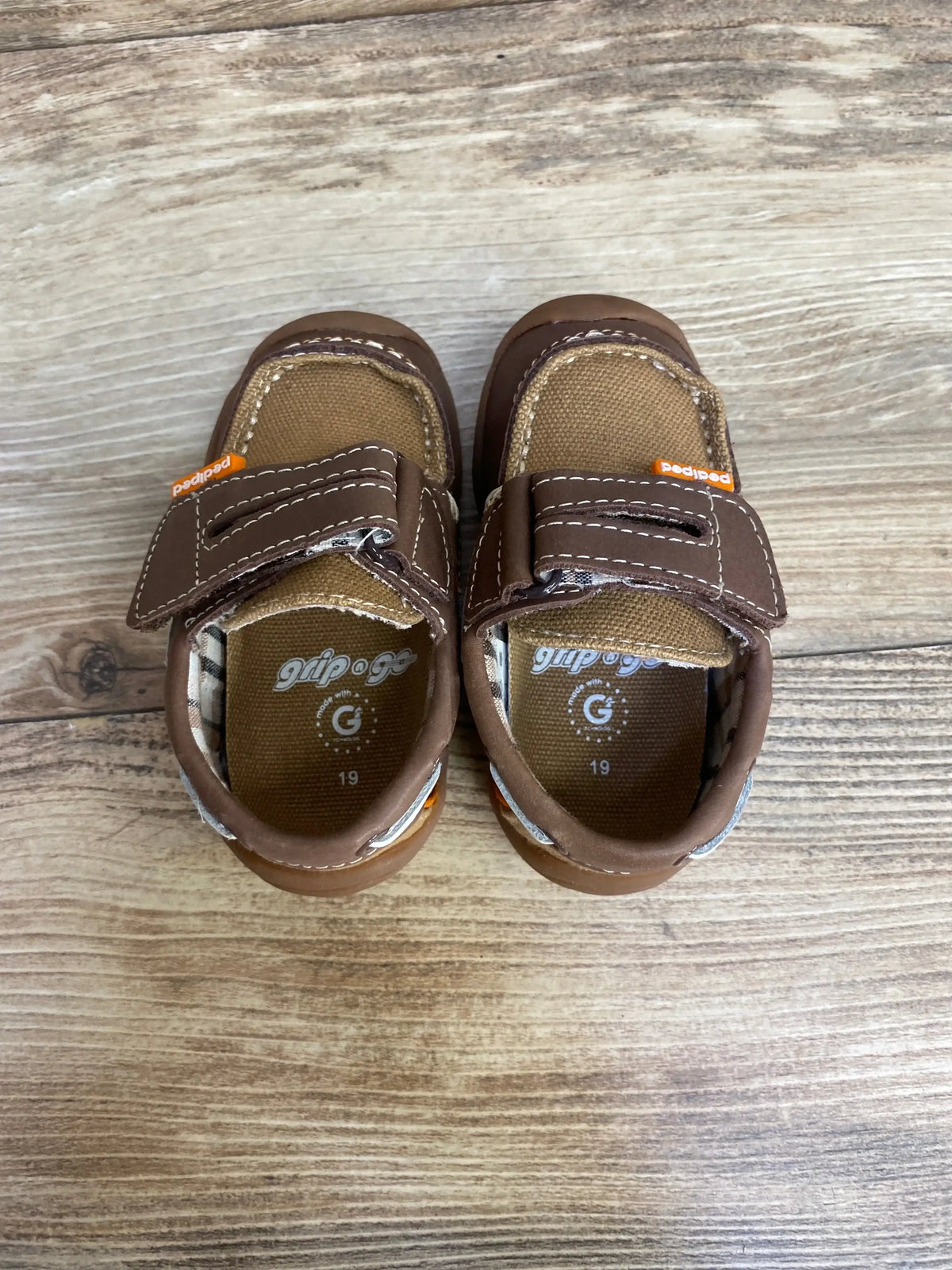 Top view of Pediped Naples brown toddler loafers with velcro, canvas upper, stitching, and orange tag on wood floor