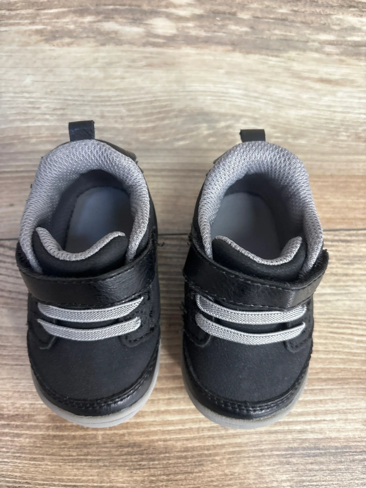 Top-down view of Stride Rite Taye 2.0 toddler sneakers in black and gray showing hook-and-loop strap, elastic laces and padded collar on wood