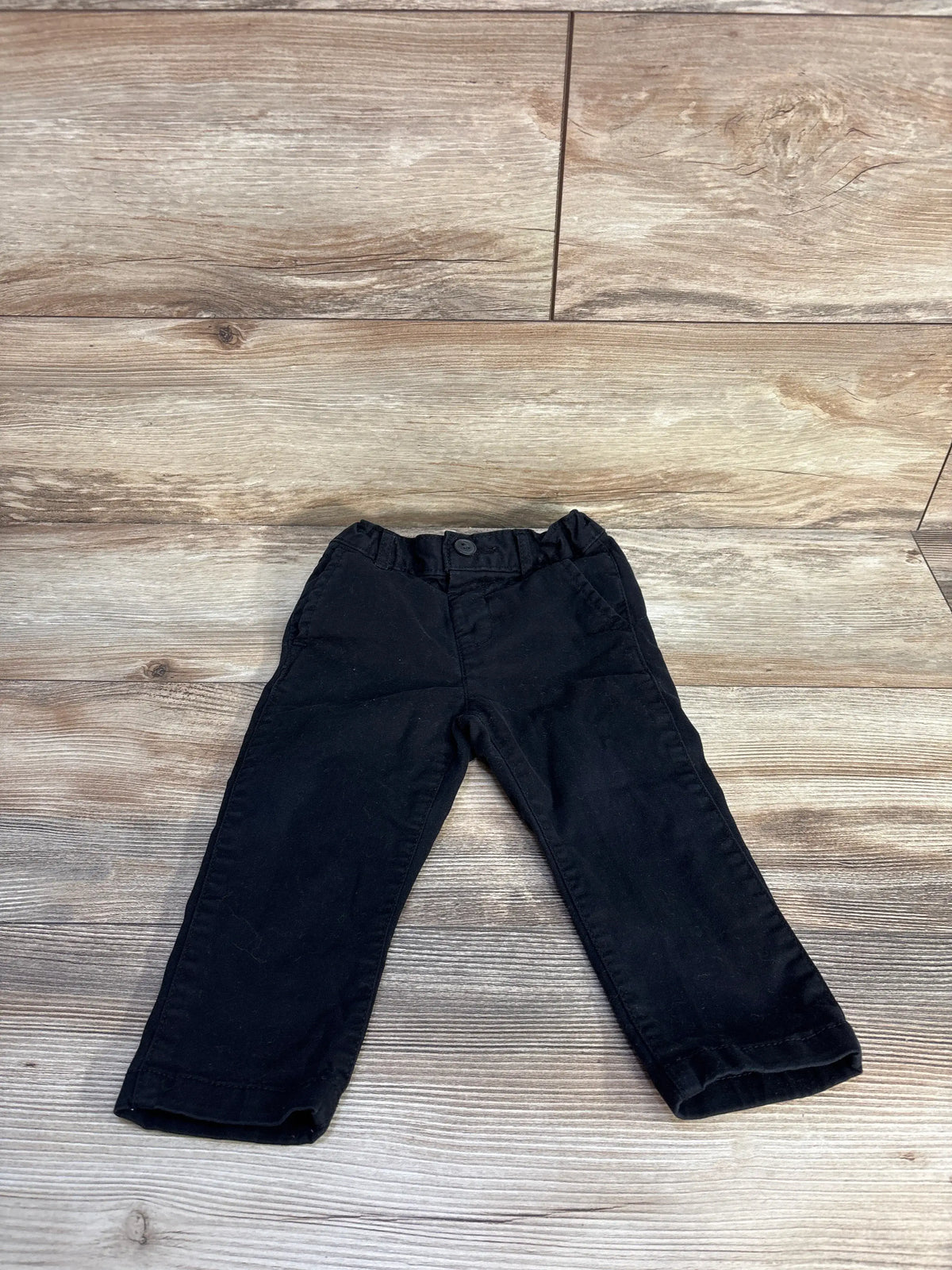 Front view of Children’s Place black pants in size 18 to 24 months showcasing button waistband and front pockets on a wood look floor