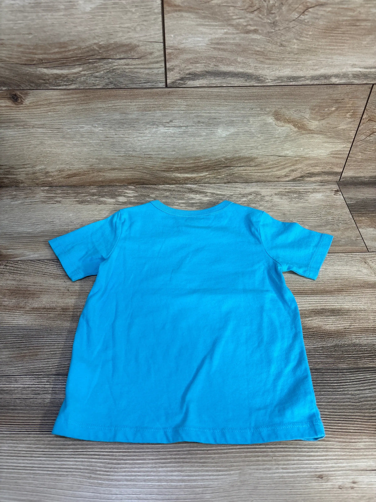 Front view of a blue Children’s Place toddler shirt showcasing short sleeves and relaxed fit on a wood-grain floor