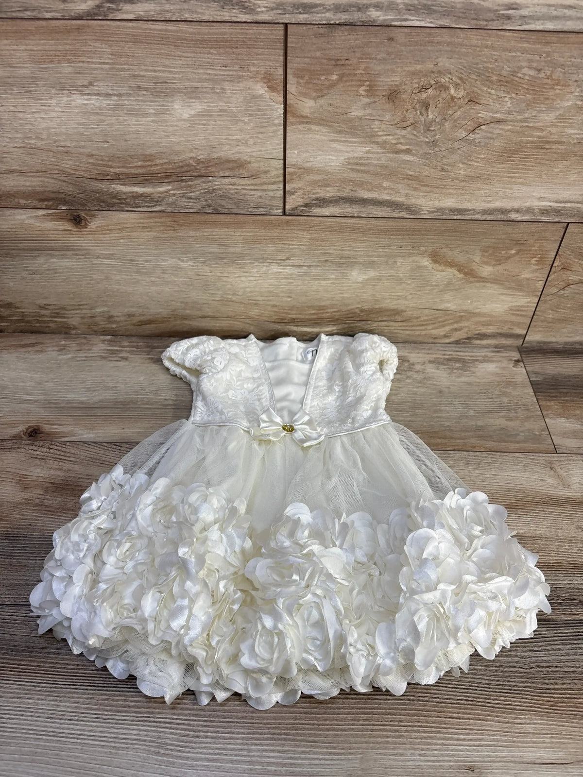 Flat lay of white Nannette lace dress with puff sleeves, satin bow with gold button, and rosette tulle skirt on wood floor