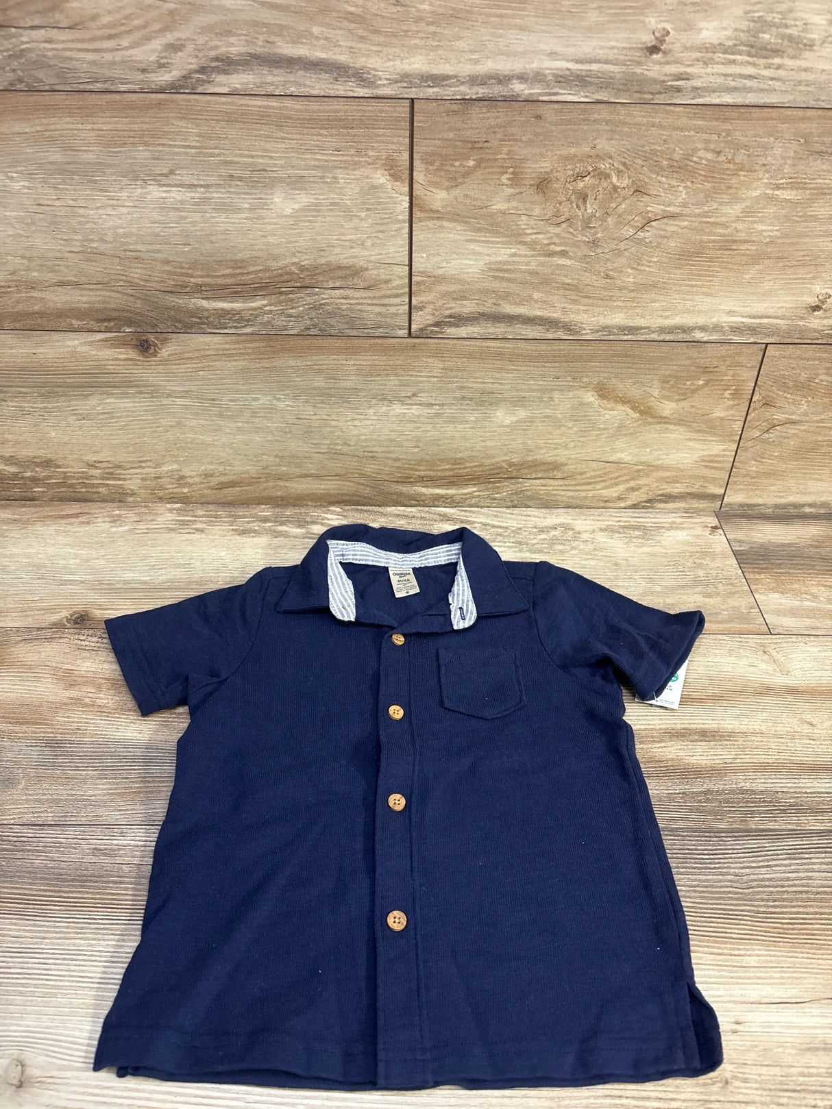 Flat-lay front of navy Oshkosh toddler button-up shirt (4T) showing wooden buttons, chest pocket, and striped inner collar on wood floor