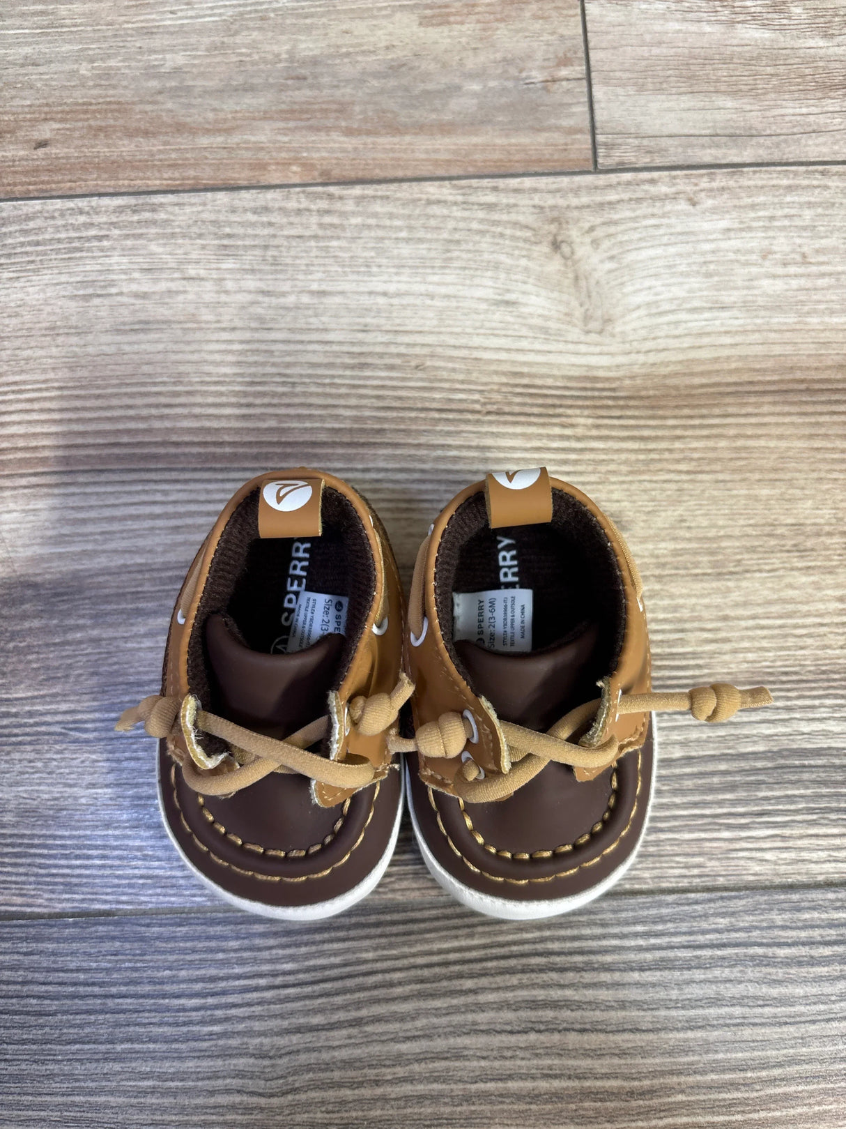Top-down view of Sperry brown and tan boat crib shoes size 2C showcasing tied laces and white rubber soles on wood floor