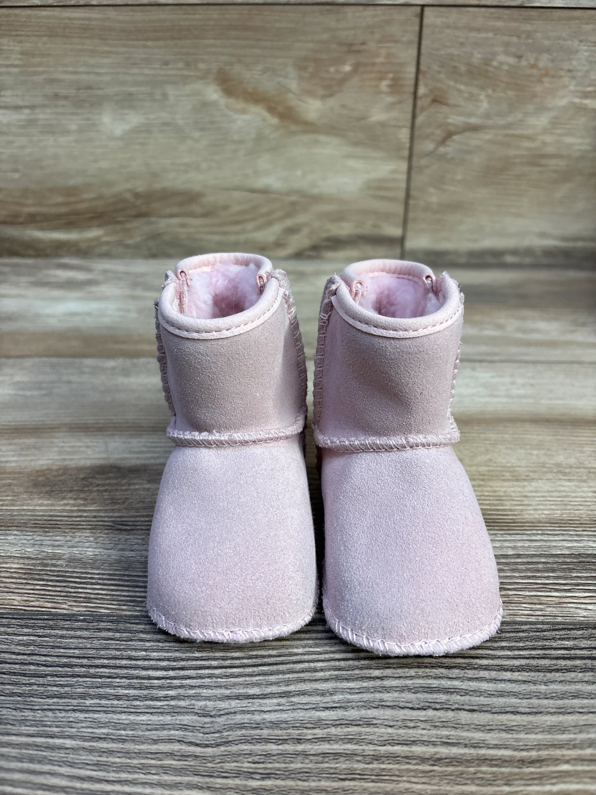 Front view of UGG Baby Classic Soft Sole baby boots in pink showing suede exterior and plush fleece lining on wood background
