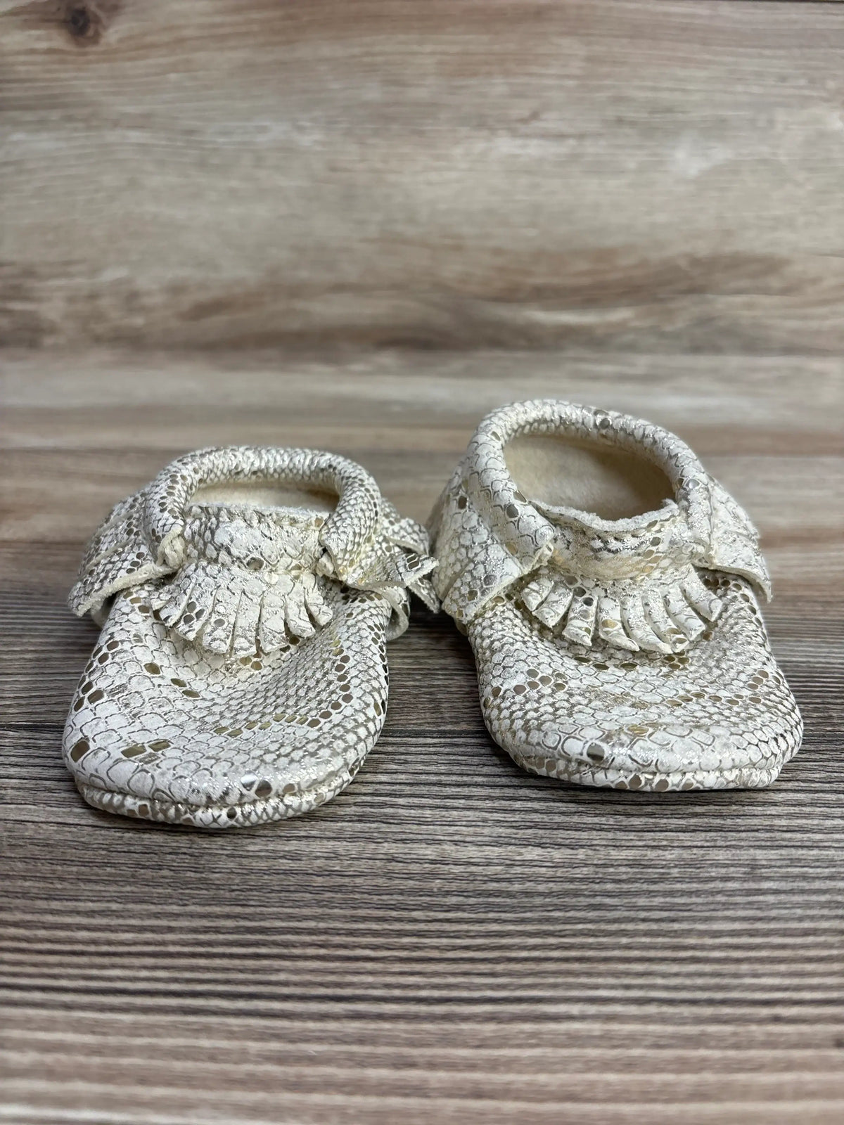 Pair of cream-colored textured baby moccasins with fringe detail on wooden surface
