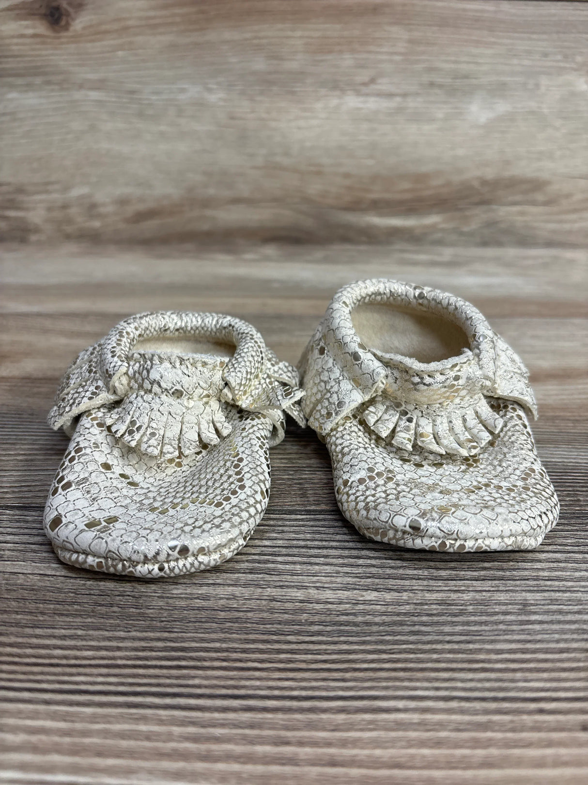 Pair of cream-colored textured baby moccasins with fringe detail on wooden surface