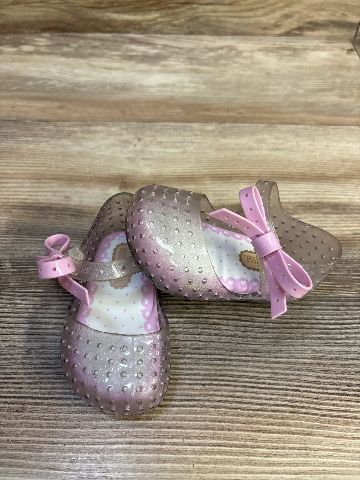Angled top view of Mini Melissa Ultragirl jelly Mary Jane shoes showcasing silver glitter finish and pink bow on wood background