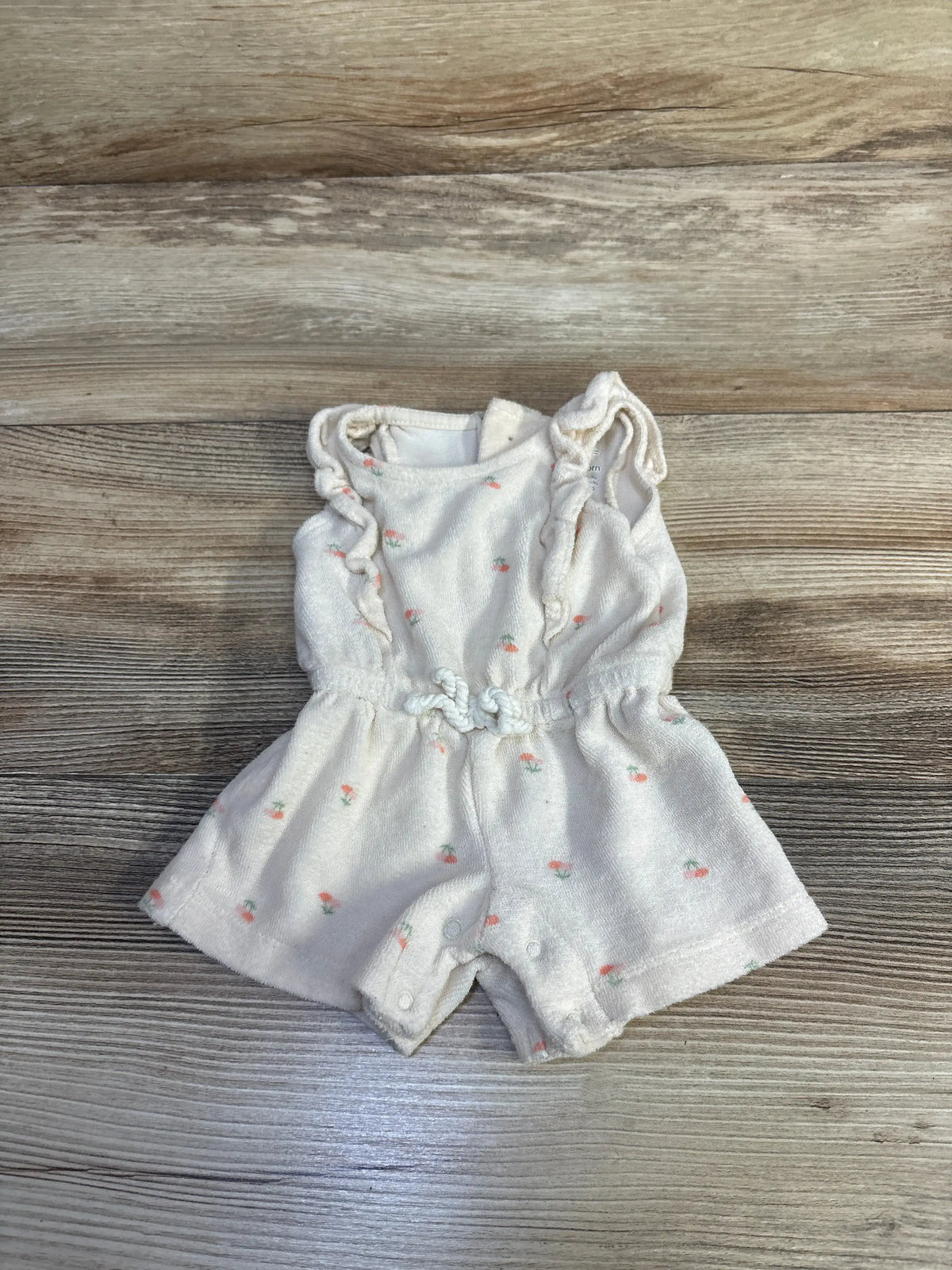 Front view of ivory floral terry cloth romper for newborns showcasing ruffled straps and snap closures on a wood background