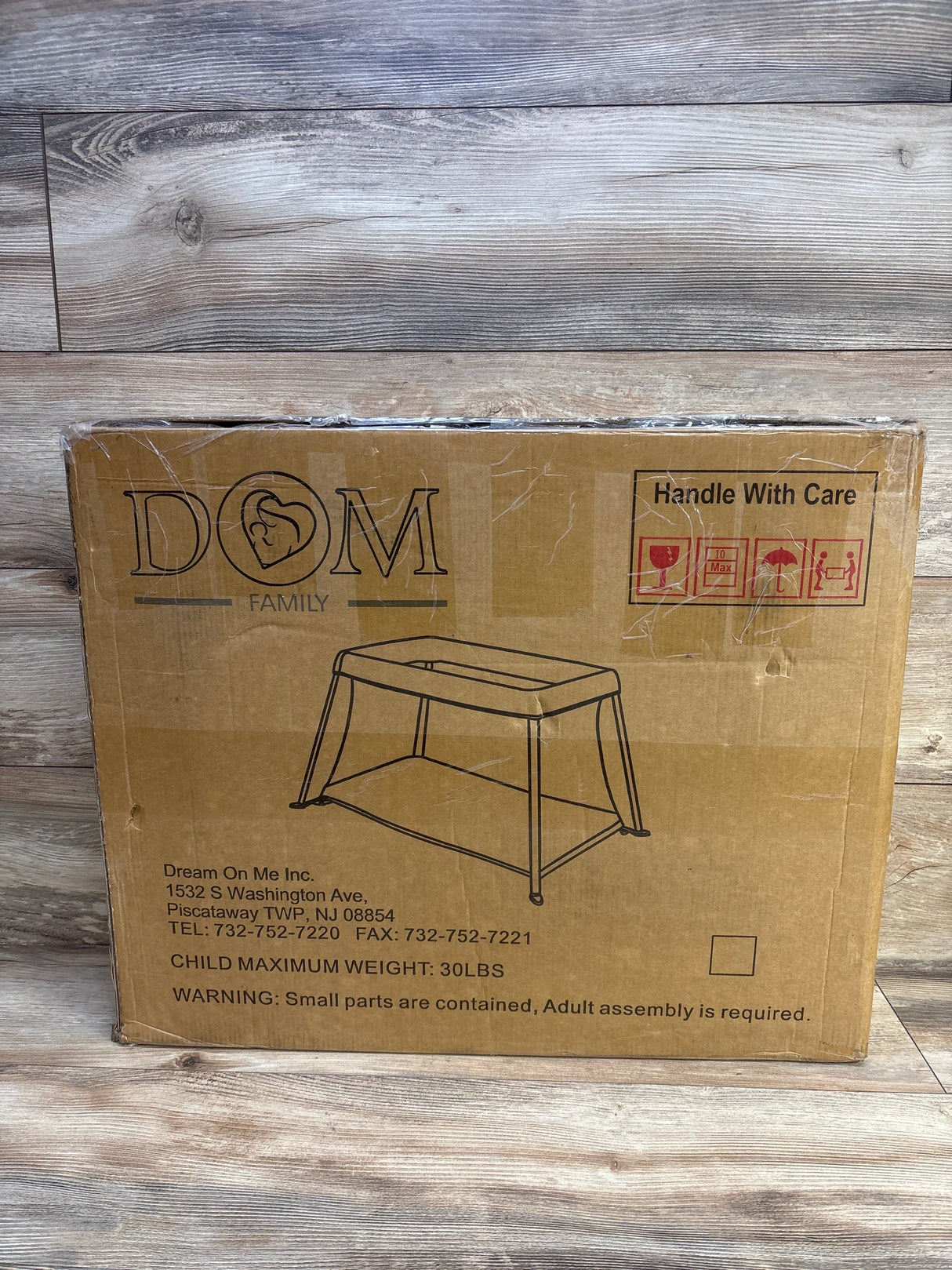 Front view of NEW Dream On Me Travel Light Playard in Blue in sealed brown DOM Family shipping box with Handle With Care label on wood plank floor