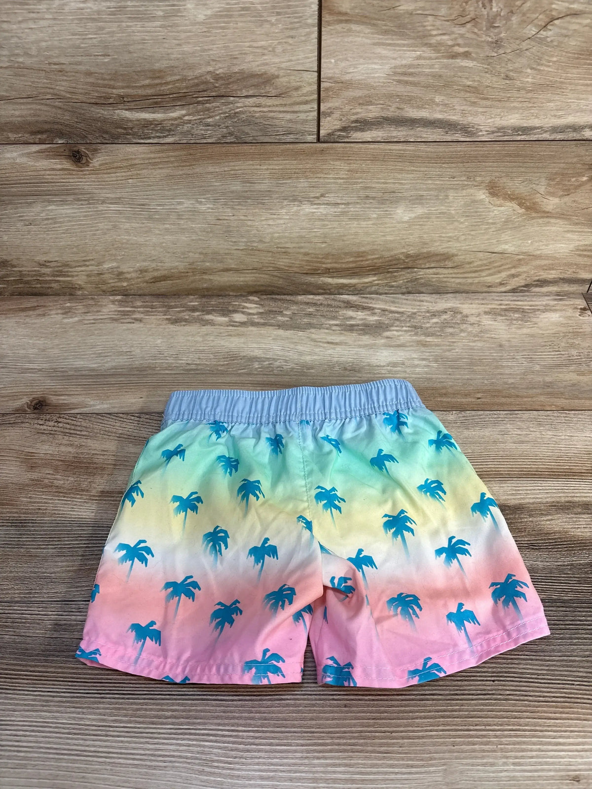 Front view of Cat & Jack Palm Tree Swim Trunks Blue sz 4T showcasing palm tree print and elastic waistband on a wood-grain background