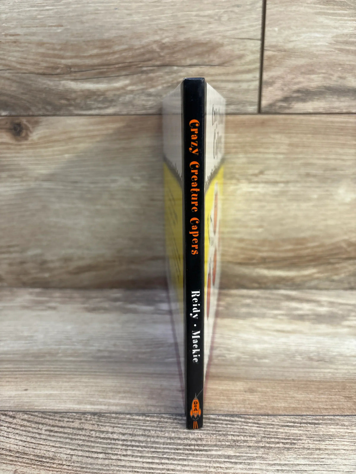 Side view of Crazy Creature Capers hardcover book showcasing its black spine with orange title lettering on a wood-grain surface