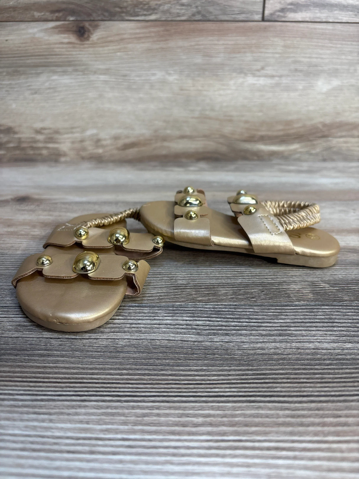 Gold toddler sandals with elastic ankle strap and decorative studs on wood background