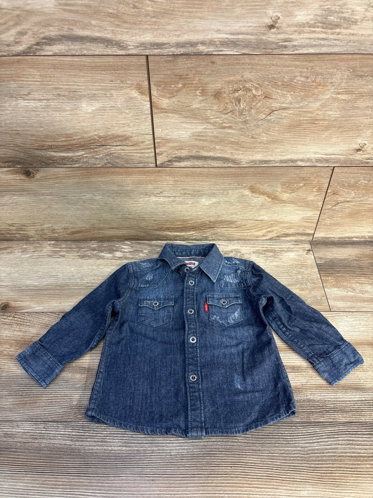 Front view of Levi’s blue chambray shirt in size 12 months showcasing long sleeves and two chest pockets on wood background