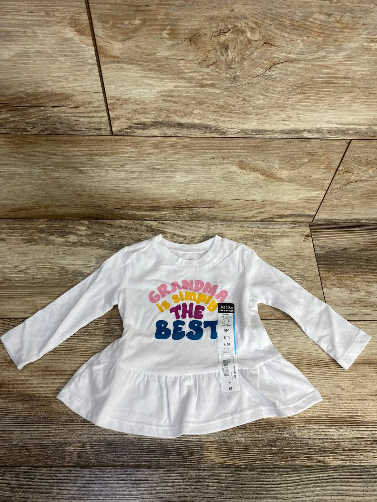 Flat lay of NEW Okie Dokie Grandma is Simply the Best peplum shirt sz 6m in white with colorful graphic and hangtag on wood plank floor