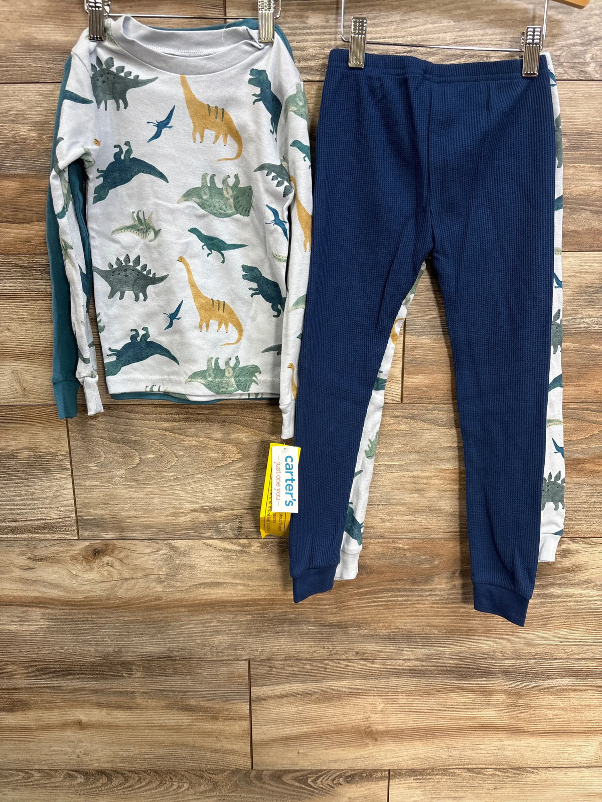 Flat lay of Just One You four-piece toddler pajama set in white and grey with dinosaur prints and textured pieces on a wood floor
