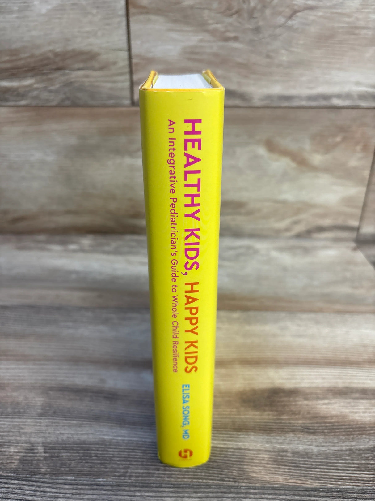 Spine view of Healthy Kids, Happy Kids hardcover showing yellow spine with vertical pink title text and author name on wood surface