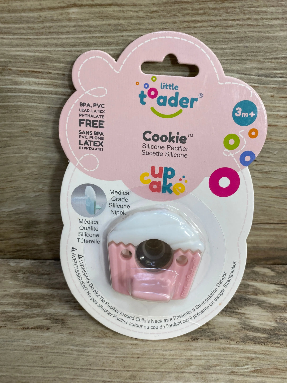 Front view of Little Toader Cookie cupcake silicone pacifier in pink on branded backing showing 3m+ age label on wood surface