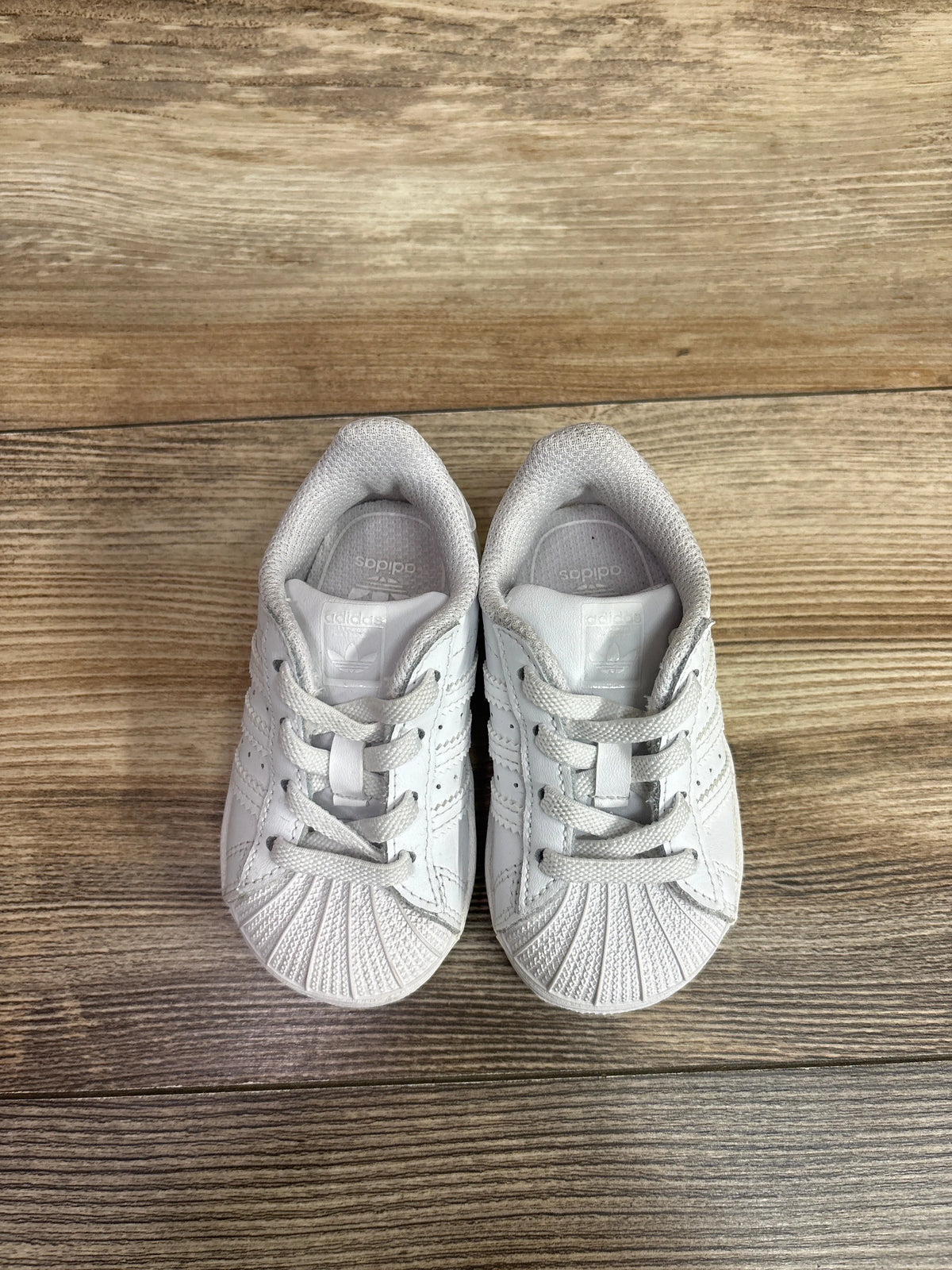 Pair of white Me n Mommy To Be sneakers for toddlers on a wooden surface.