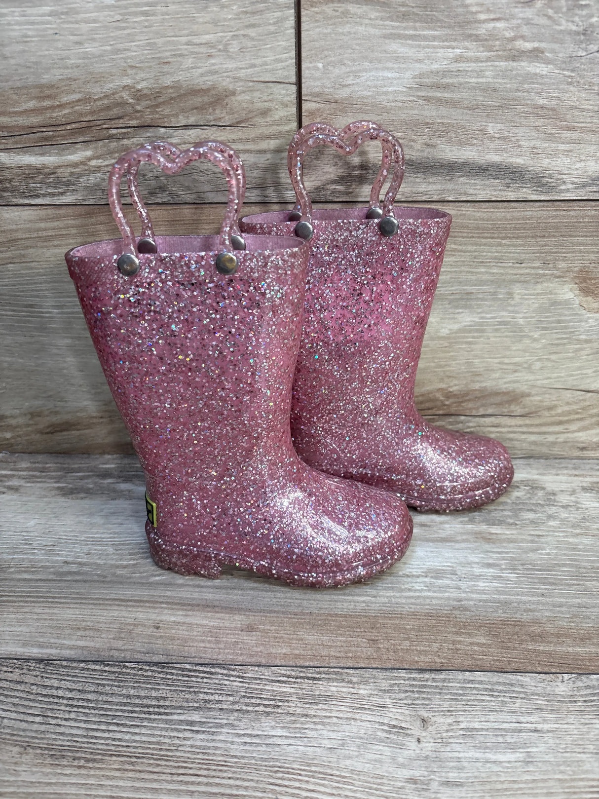 Pink glitter kids rain boots with heart-shaped handles on wood background