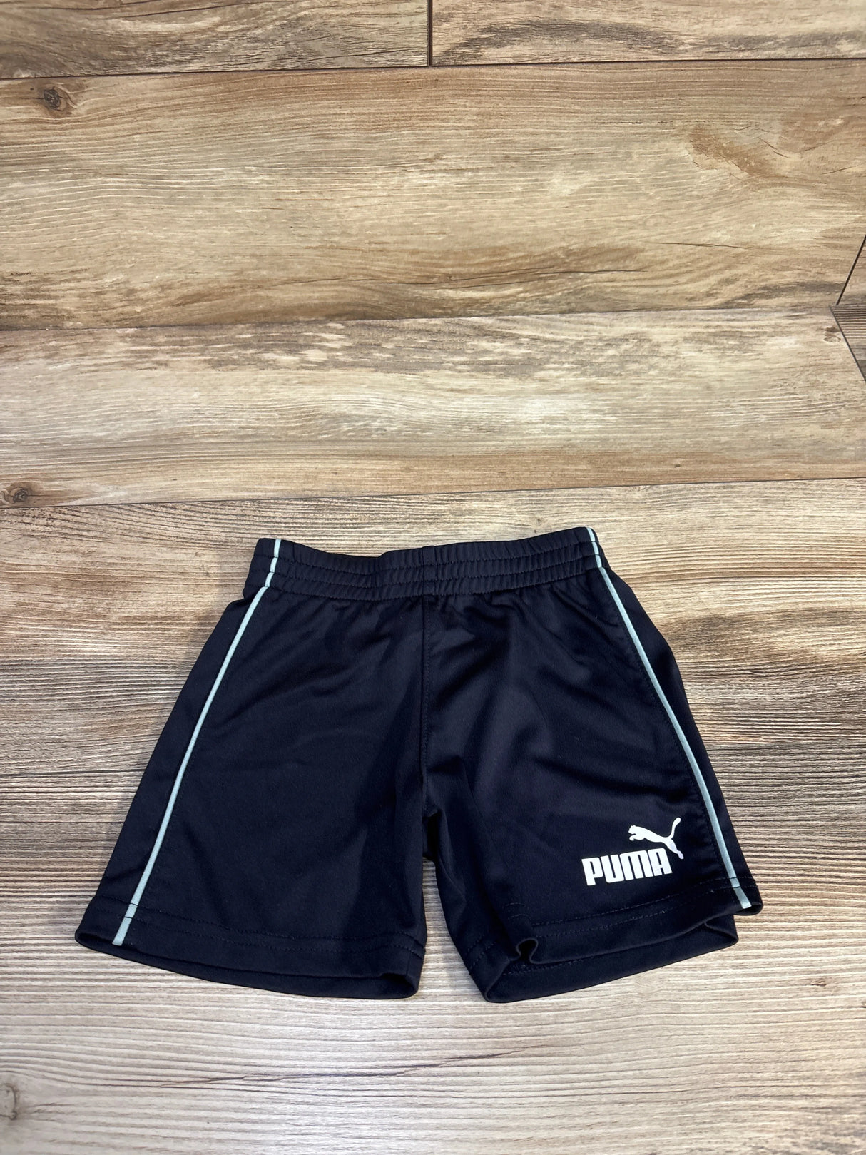 Front view of toddler black Puma active shorts showcasing elastic waistband, light blue side piping and white Puma logo with wooden floor background