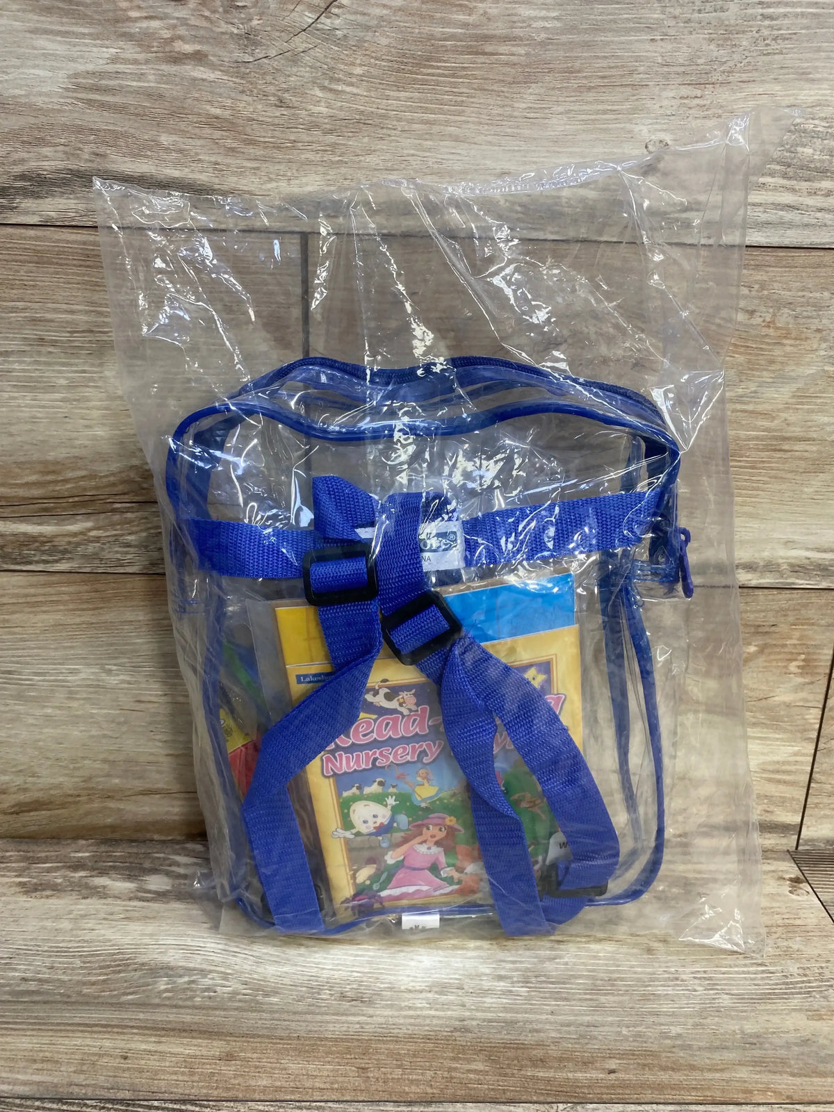 Front view of Lakeshore Ready for Preschool Backpack showcasing clear vinyl body and blue straps with nursery books inside on a wood background