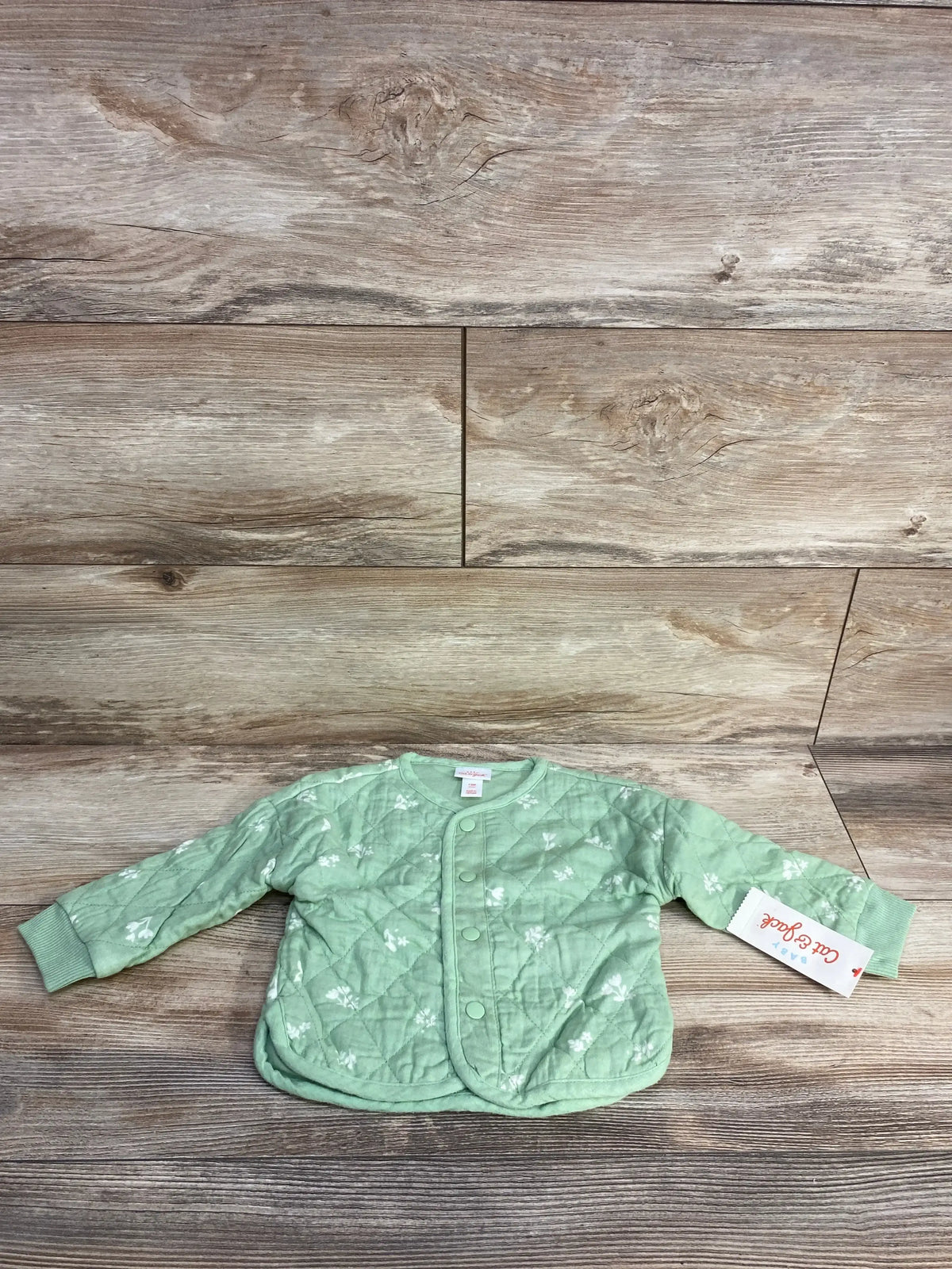 Light green quilted baby jacket with white floral patterns and snap buttons on wooden floor