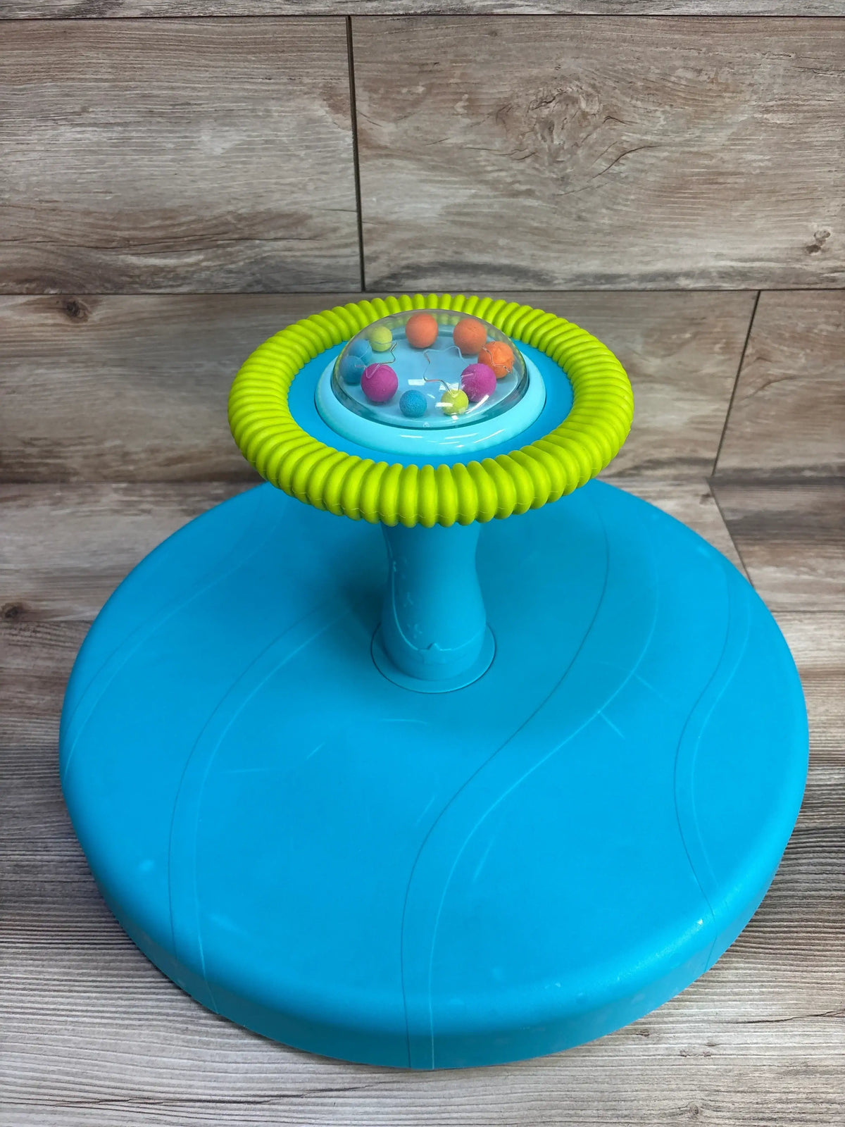 Top view of B. Toys Twirly Time spinning toy showing clear dome with colorful balls and green ring on wood backdrop